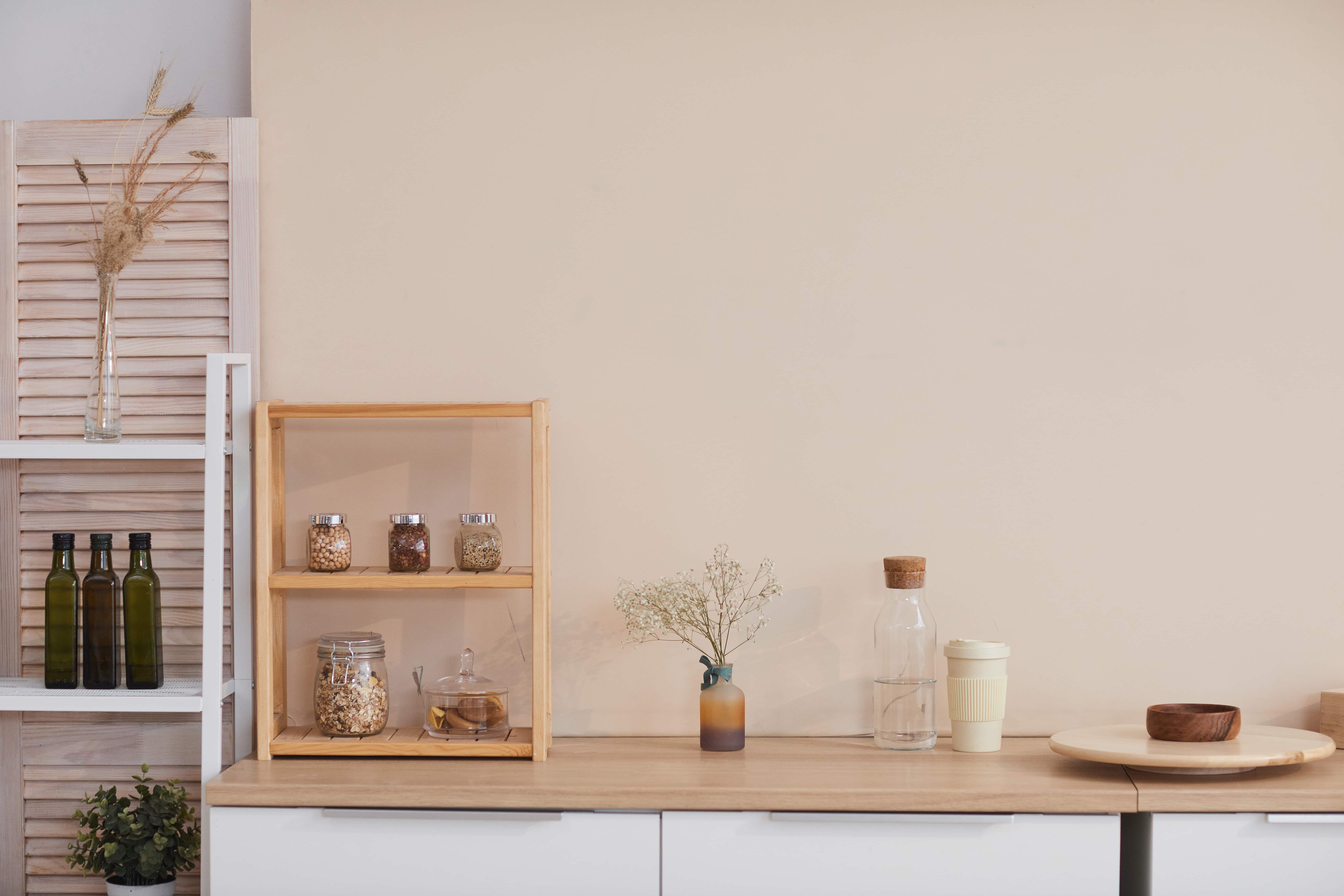 Es ist eine Küchenzeile mit weißen Fronten und einer Arbeitsplatte in Holzoptik zu sehen. Daneben steht ein offenes, weißes Regal. Auf der Arbeitsfläche stehen ein kleines Holzregal sowie mehrere Dekoelemente. Dahinter ist eine beigefarbene Wand zu sehen.