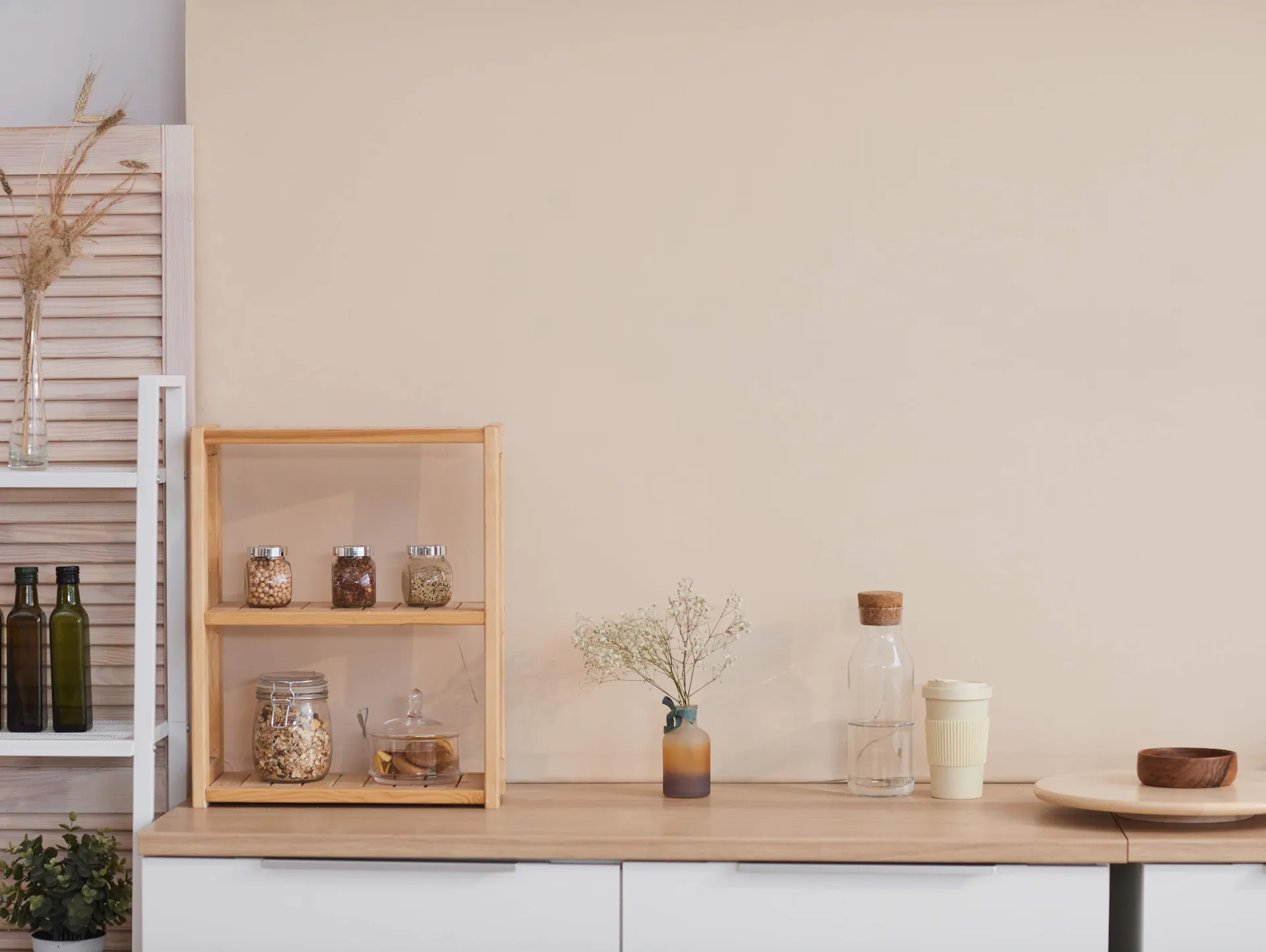 Es ist eine Küchenzeile mit weißen Fronten und einer Arbeitsplatte in Holzoptik zu sehen. Daneben steht ein offenes, weißes Regal. Auf der Arbeitsfläche stehen ein kleines Holzregal sowie mehrere Dekoelemente. Dahinter ist eine beigefarbene Wand zu sehen.