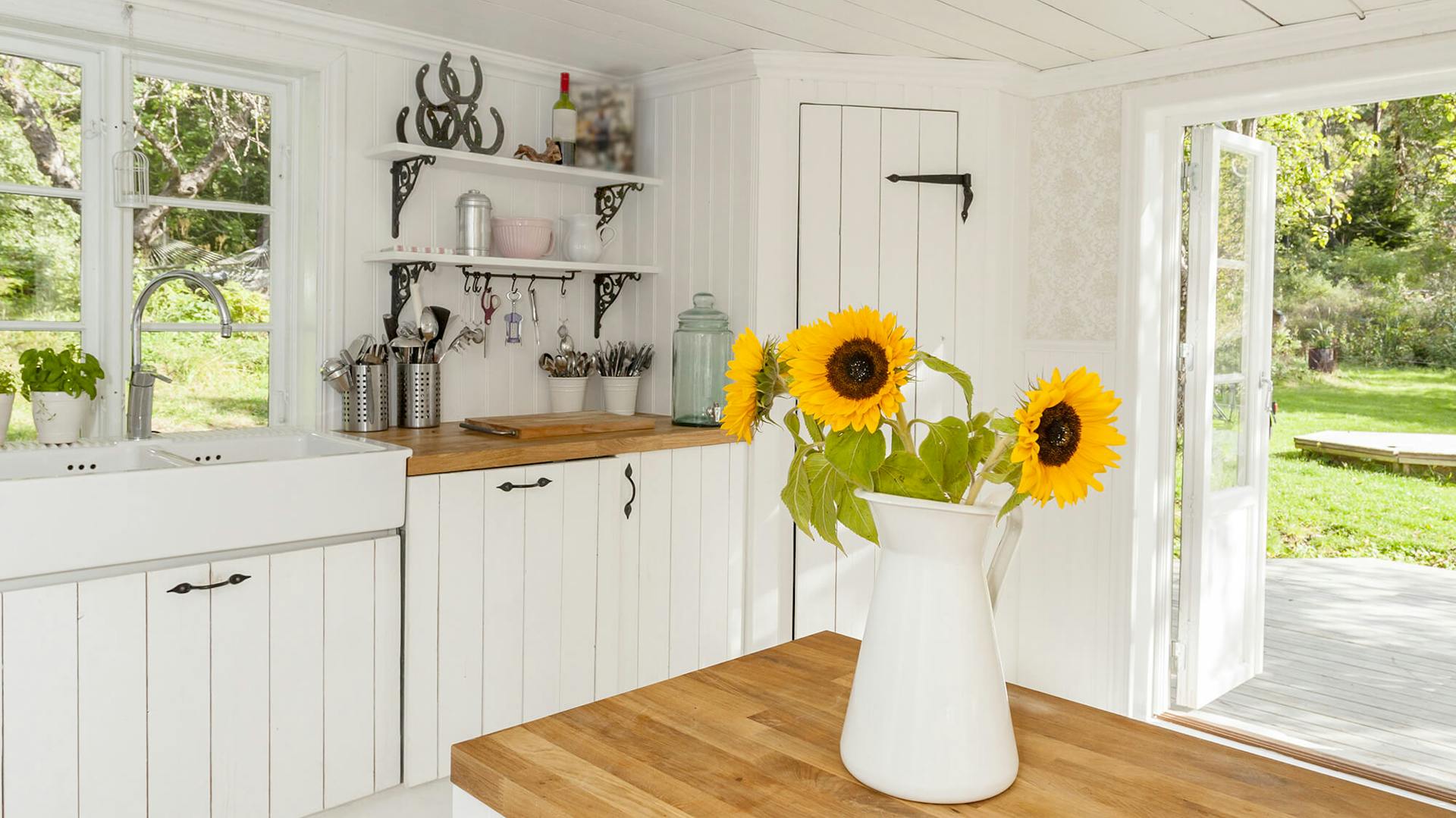 Weiße Landhausküche. Vorne steht ein hölzerner Tisch mit einer Vase mit Sonnenblumen.