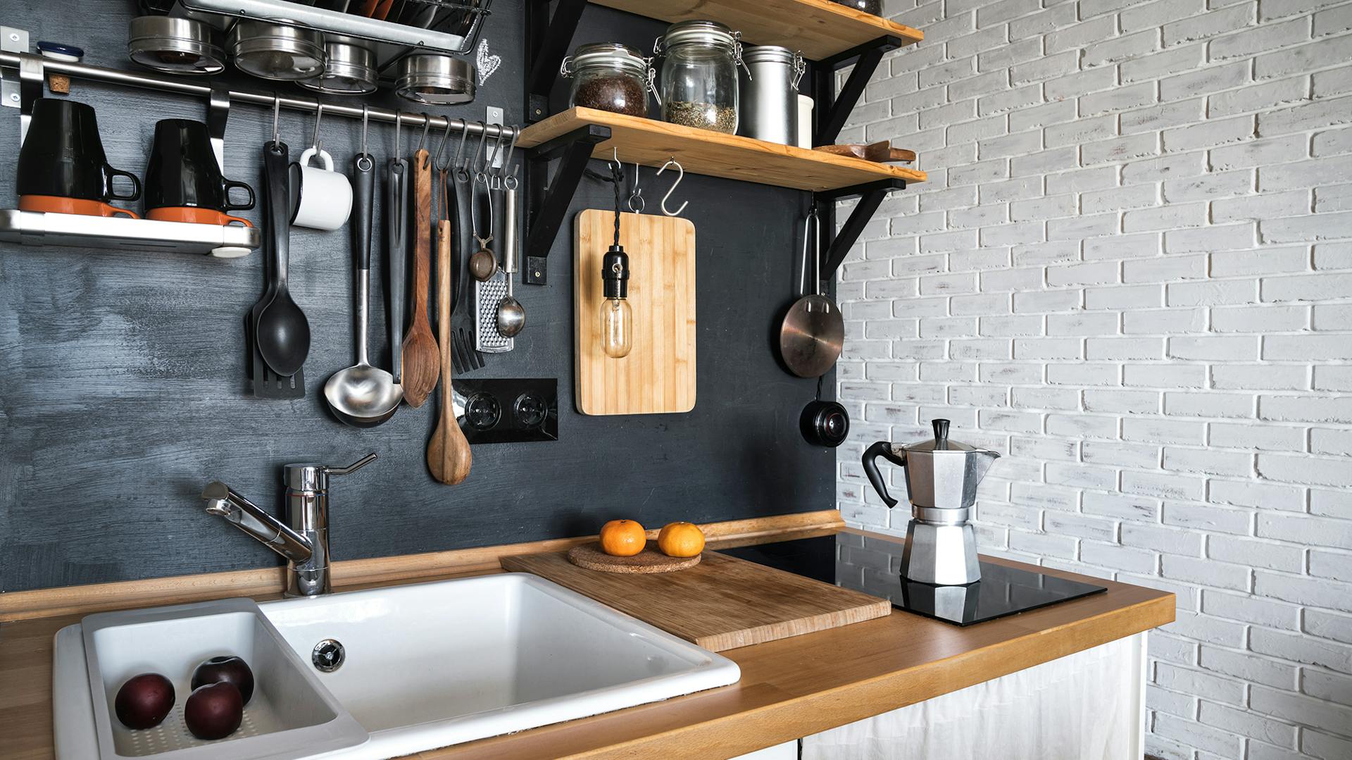 Weiße Küchenzeile mit Holzarbeitsfläche. Darauf ein weißes Keramikwaschbecken und ein Kochfeld. Die Wand im Hintergrund ist schwarz. An der hängen Wandregale und Küchenutensilien.