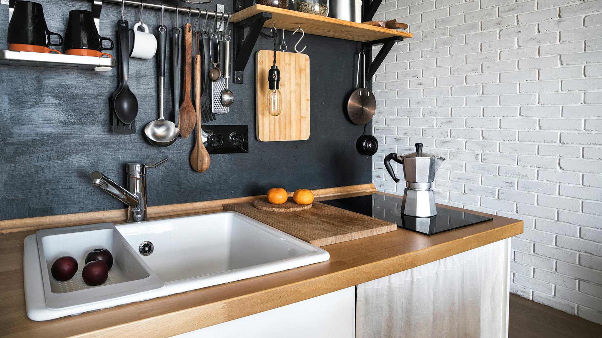 Weiße Küchenzeile mit Holzarbeitsfläche. Darauf ein weißes Keramikwaschbecken und ein Kochfeld. Die Wand im Hintergrund ist schwarz. An der Wand hängen Wandregale und Küchenutensilien.