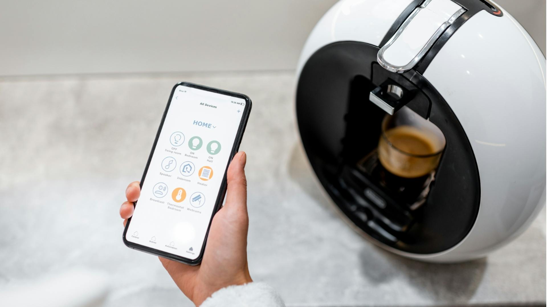 Person bedient Kaffeemaschine mit dem Smartphone. Im Hintergrund läuft Kaffee aus Maschine in Tasse.