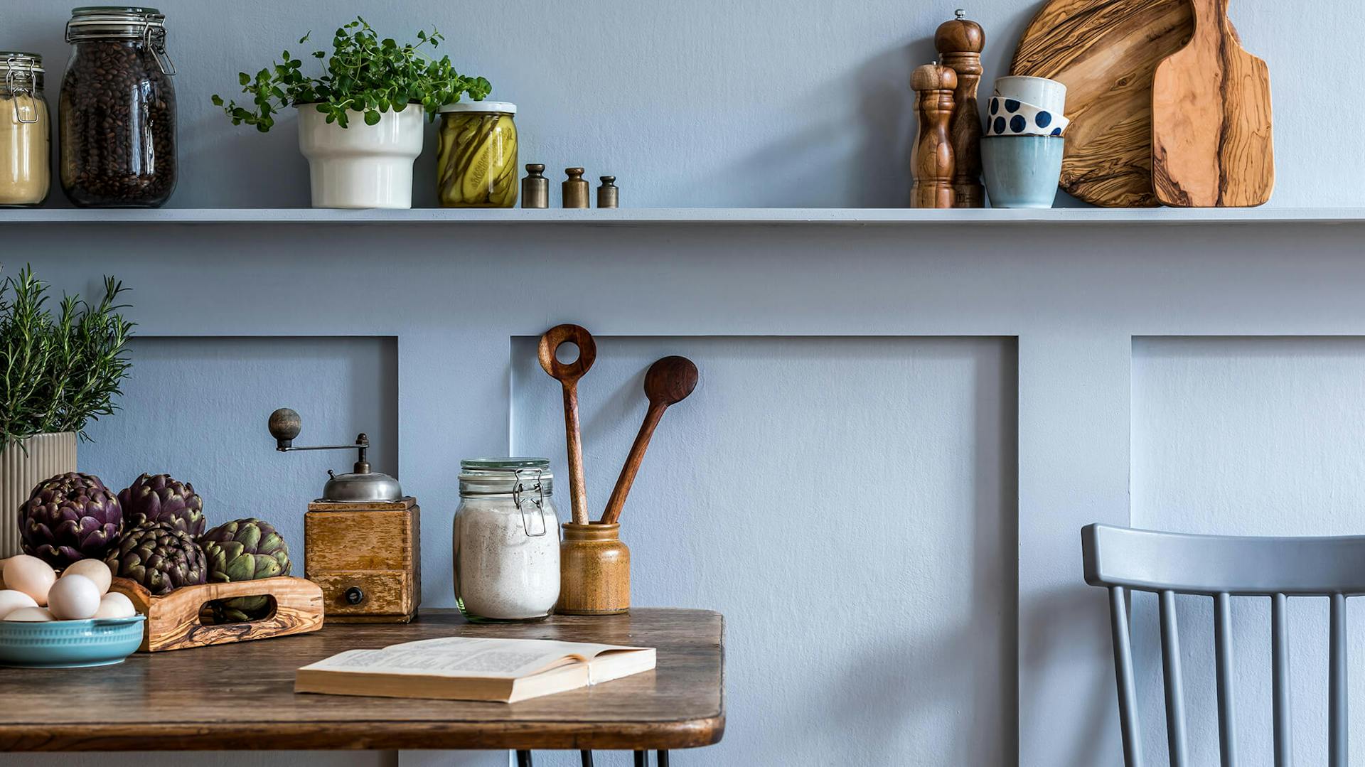 Rustikaler Küchentisch vor eisblauer Wand. Auf einem Wandregal stehen rustikale Küchenaccessoires und Kräuter