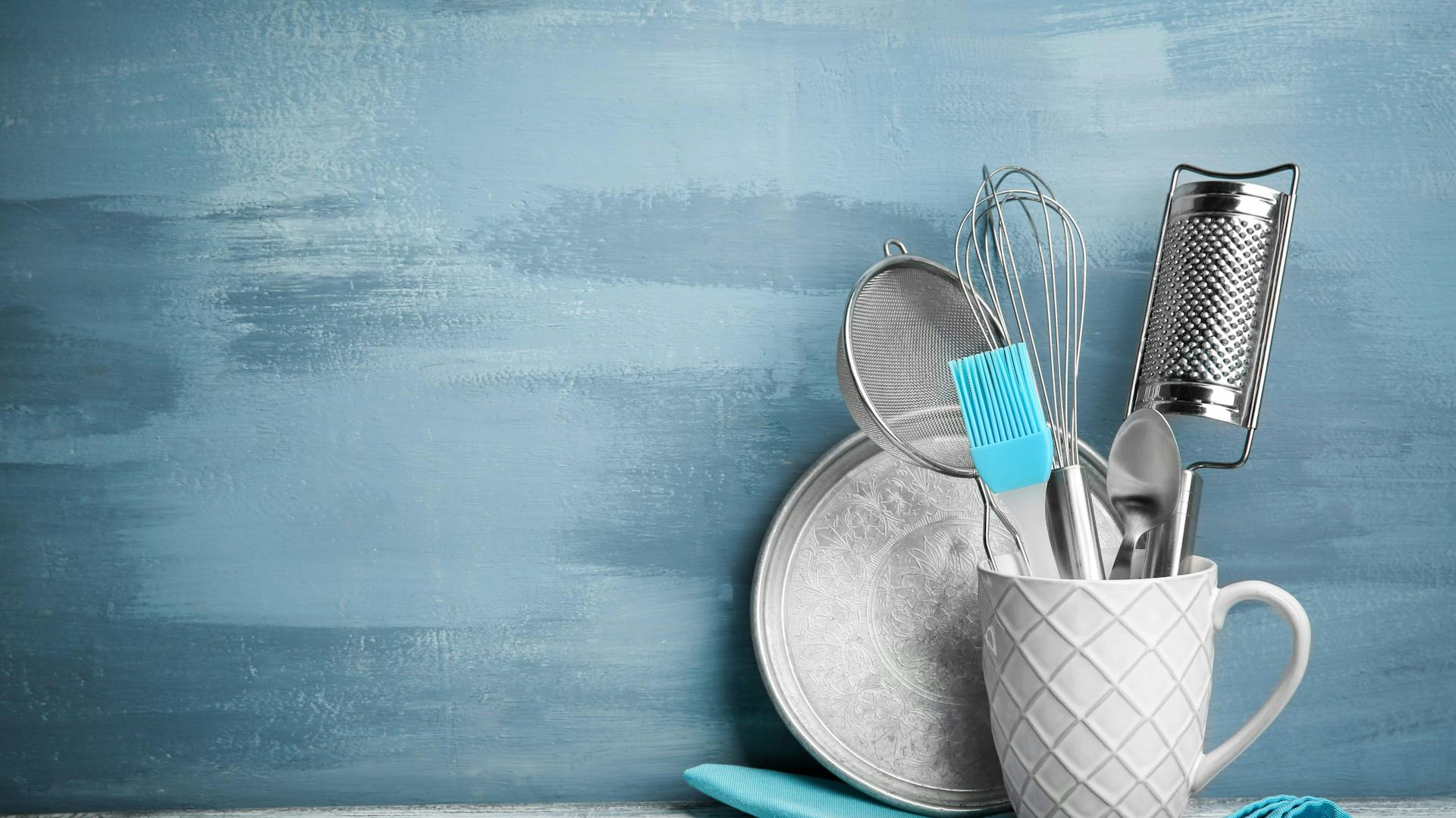 Küchenutensilien in einer weißen Tasse, platziert auf einem blauen Geschirrhandtuch auf einer grauen Arbeitsplatte vor blauer Küchenwand.