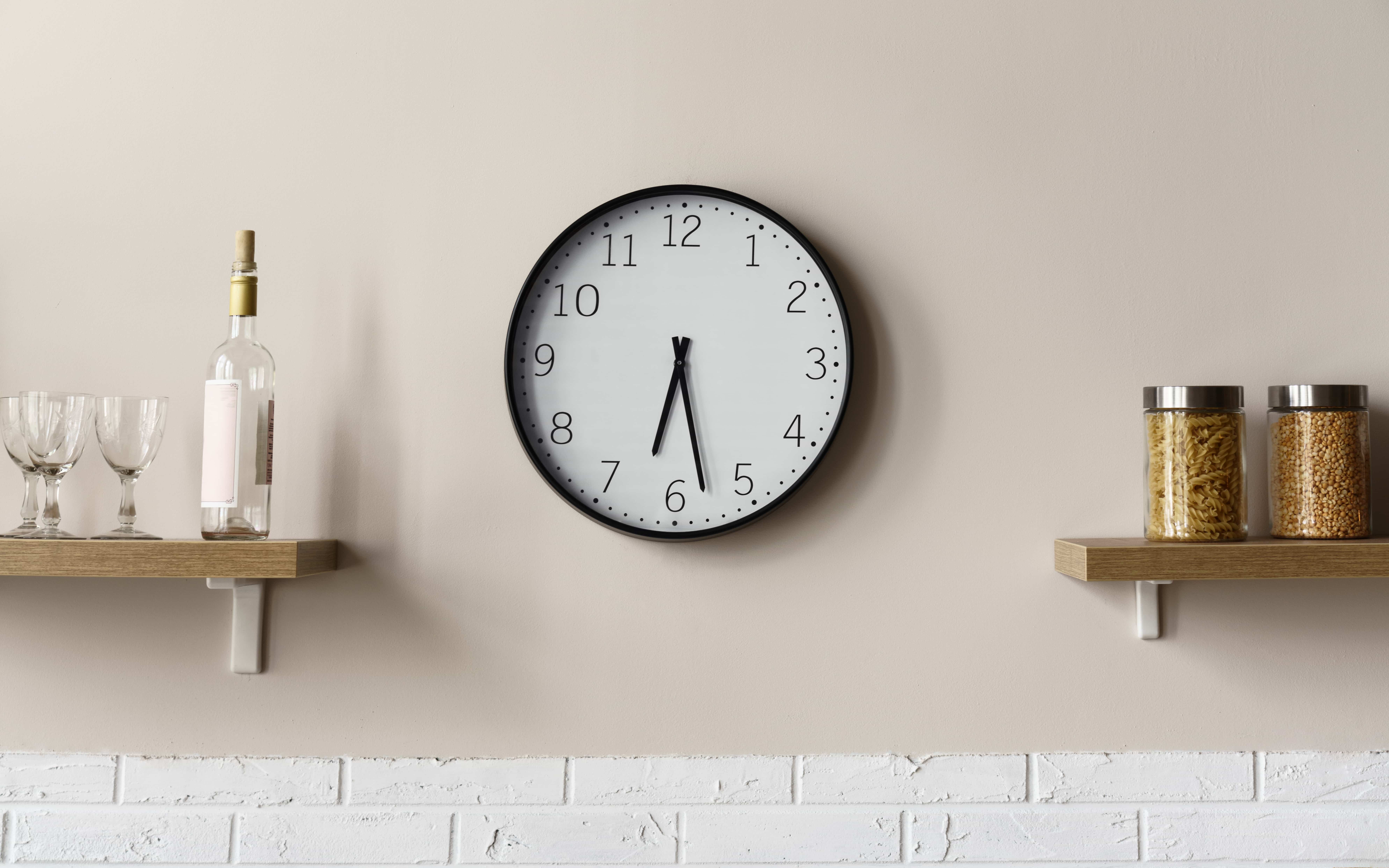 Schwarz-weiße Küchenuhr an einer beigen Küchenwand zwischen zwei Regalen mit Küchenutensilien.