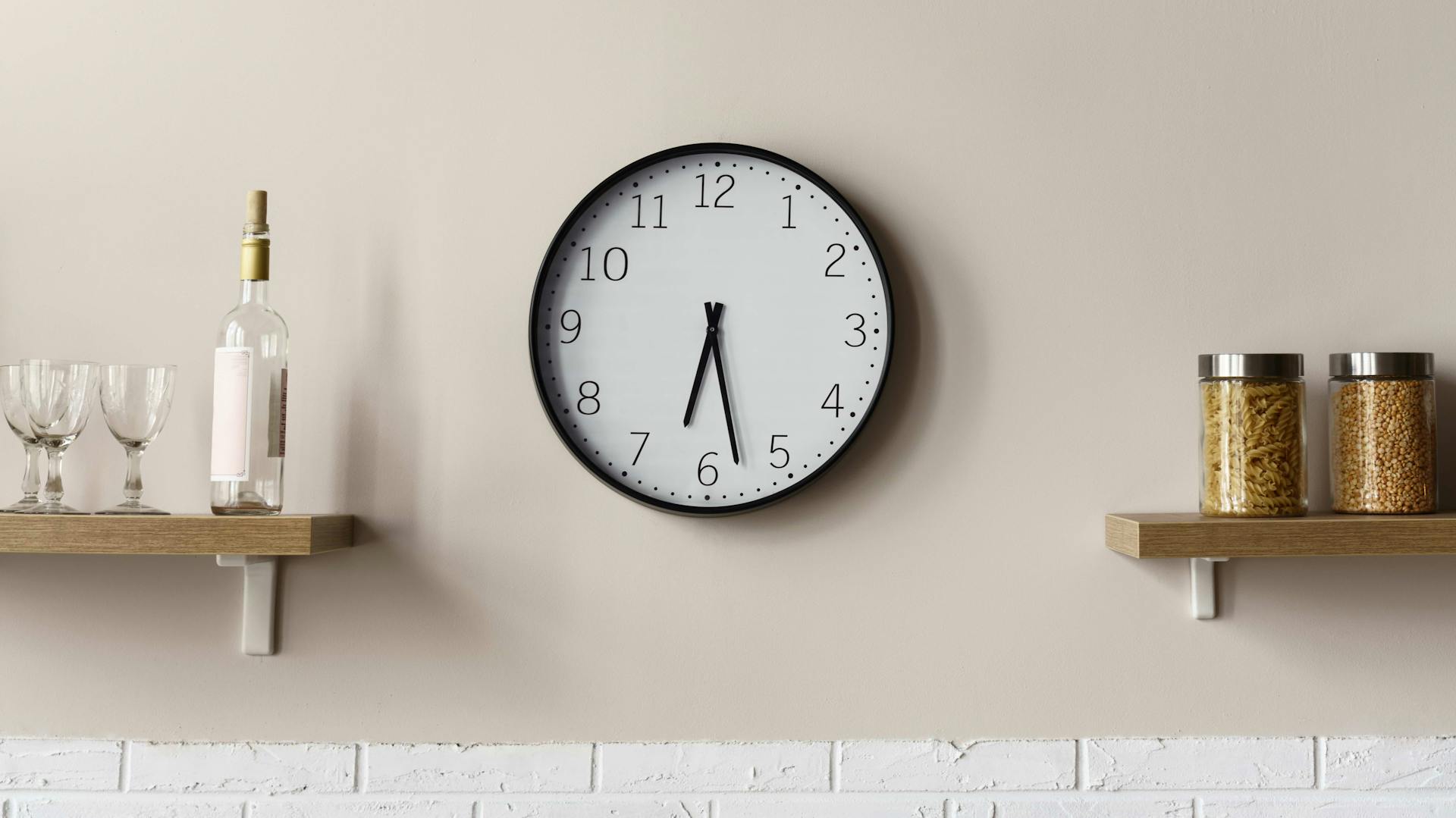 Schwarz-weiße Küchenuhr an einer beigen Küchenwand zwischen zwei Regalen mit Küchenutensilien.