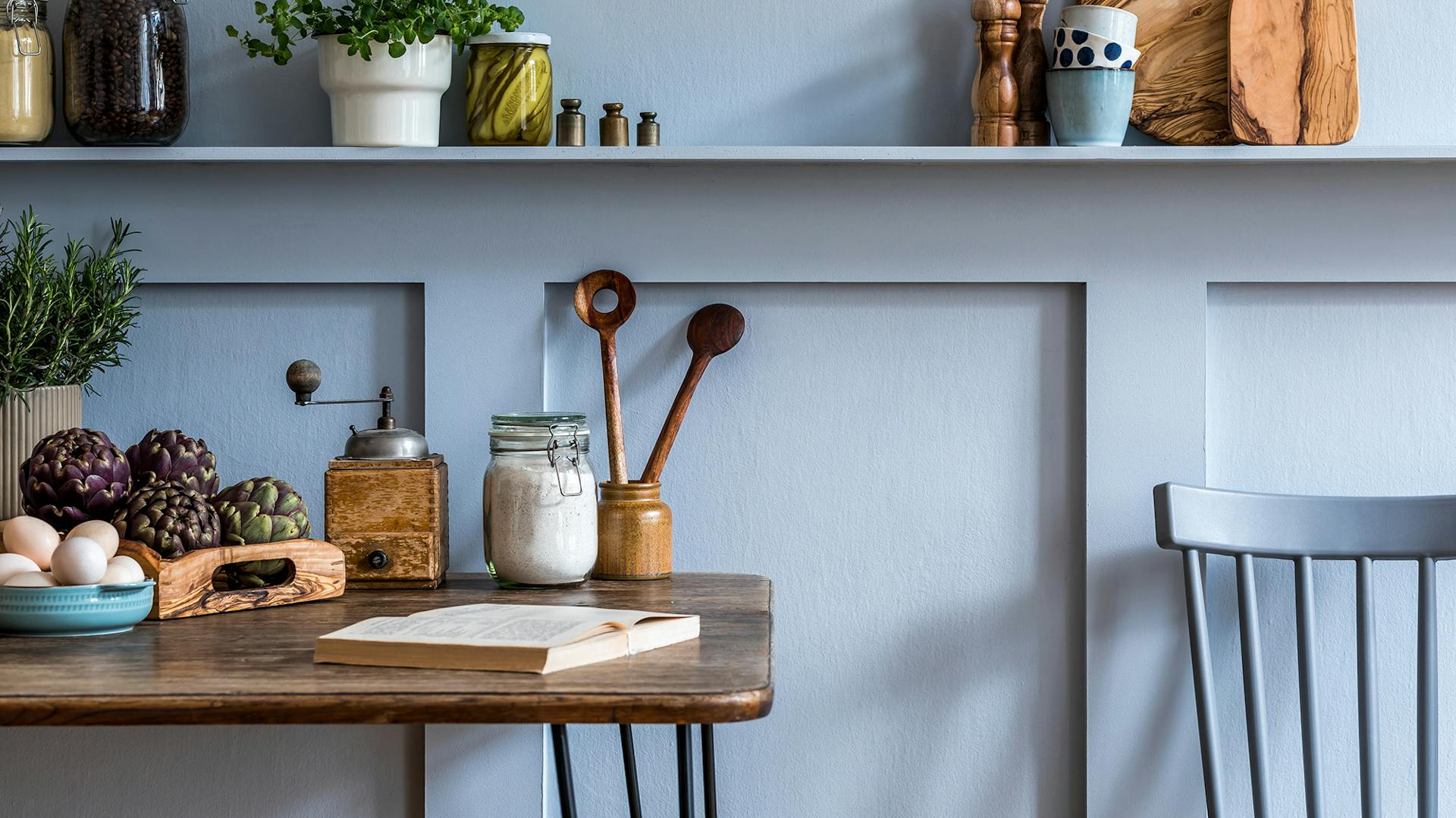 Küchentisch, Küchenstuhl und diverse Küchenutensilien vor einer hellblauen Wand mit Vertäfelung.