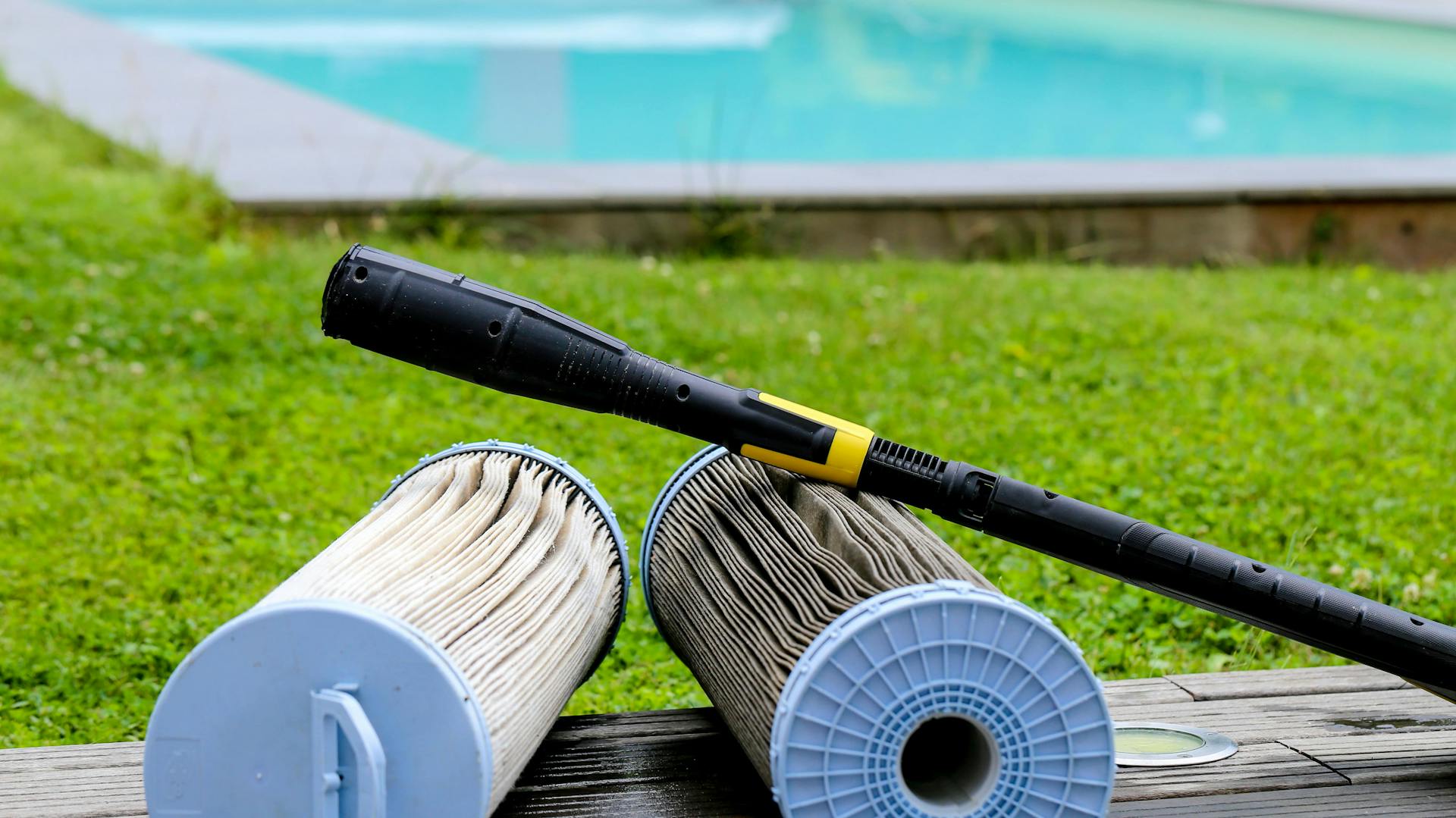 Un filtro a cartuccia per piscina sporco e uno pulito sono davanti a una piscina con un’idropulitrice ad alta pressione.