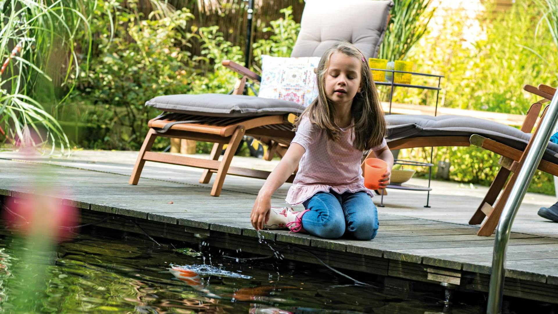 Ein Kind sitzt am Ufer des Schwimmteichs mit Liegestühlen im Hintergrund.