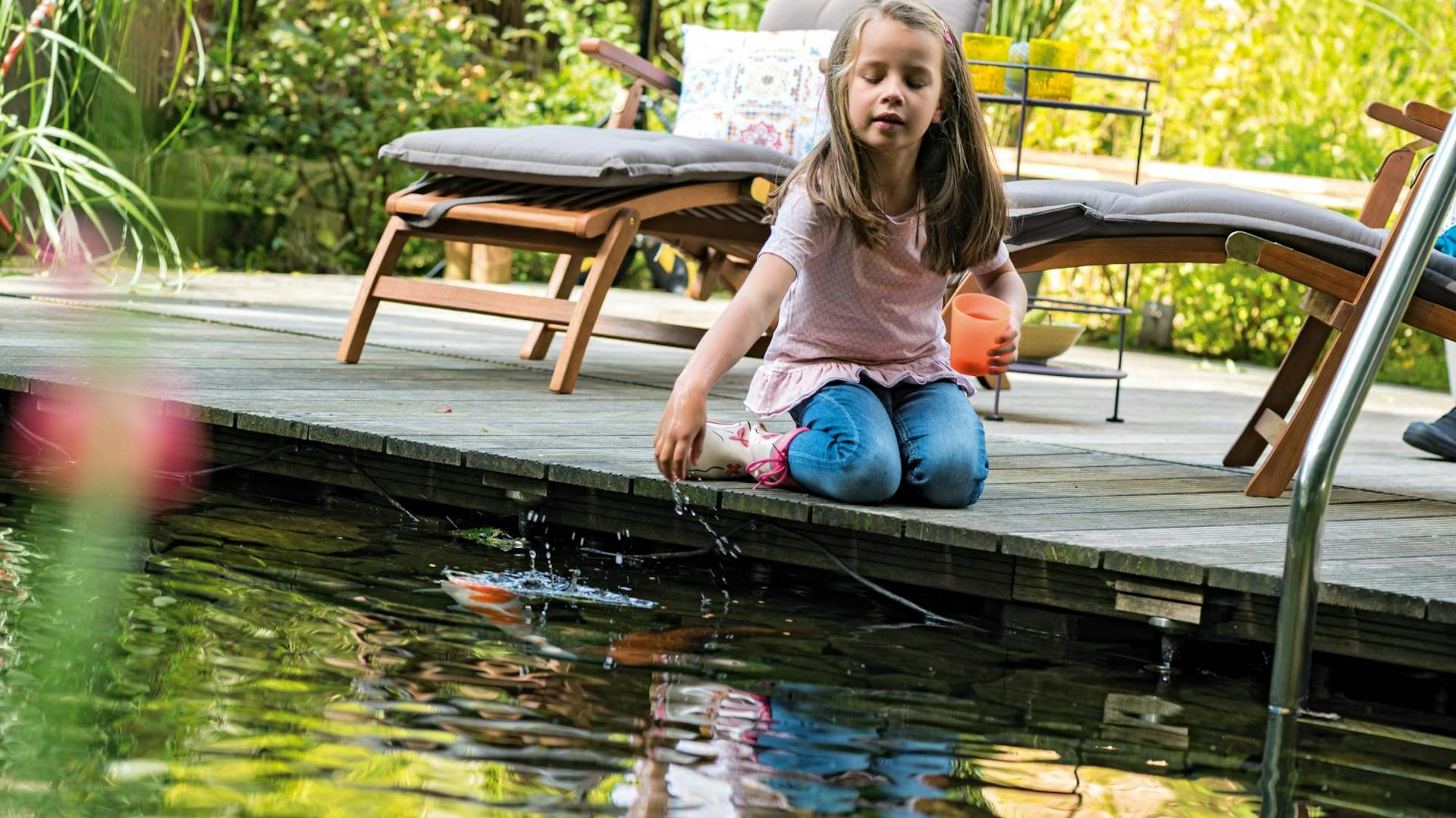 Ein Kind sitzt am Ufer des Schwimmteichs mit Liegestühlen im Hintergrund.