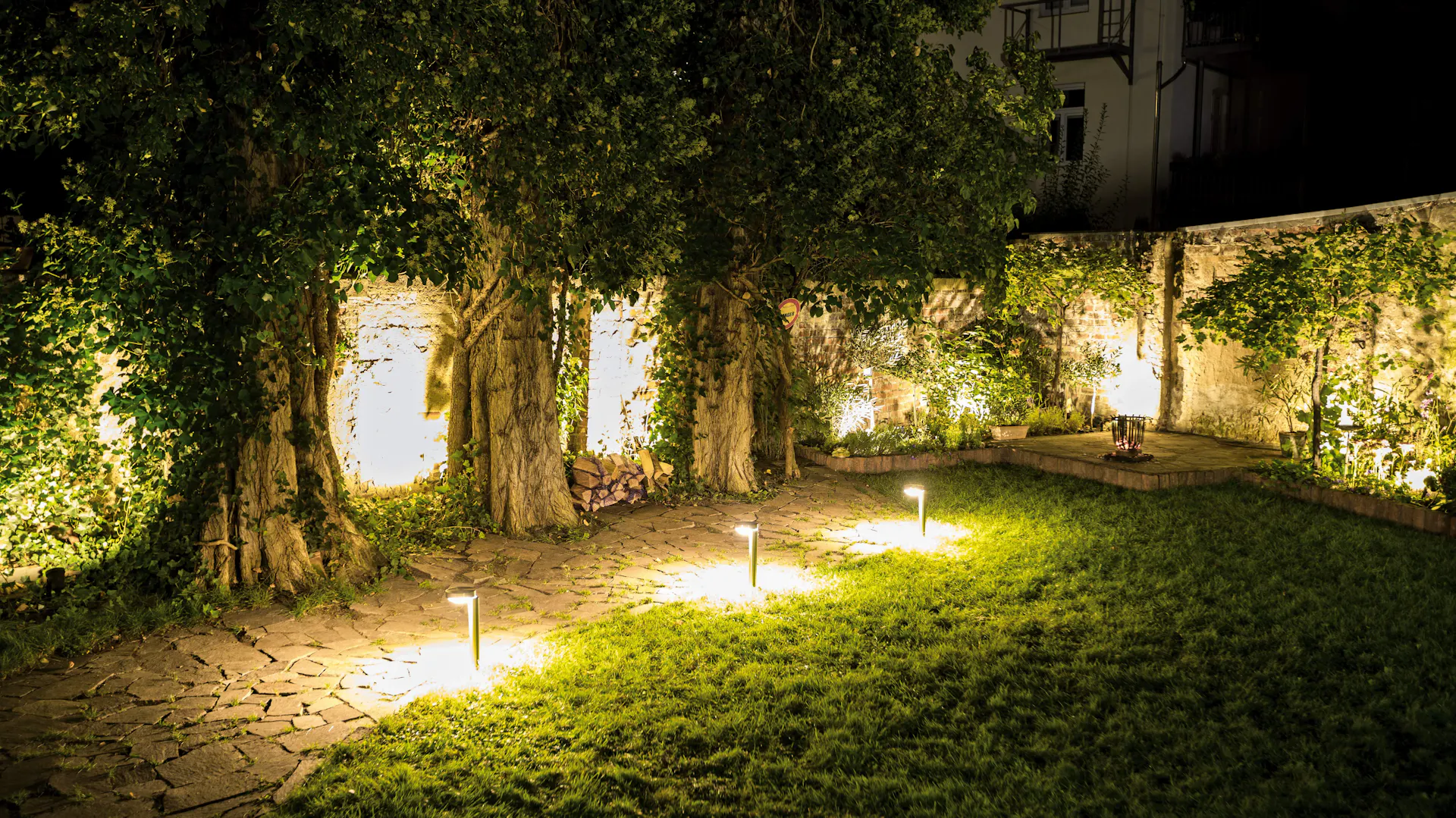 Kültéri lámpák a kertben pázsittal, térkővel, fákkal és fallal éjszaka