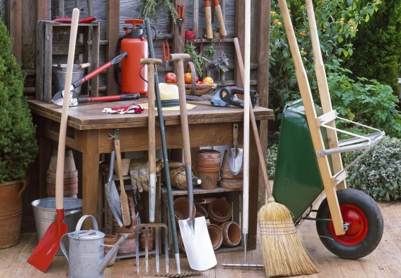 Különböző kerti szerszámok gyűjteménye a fából készült munkapad előtt, köztük sövényvágó, ásó, seprű és talicska.