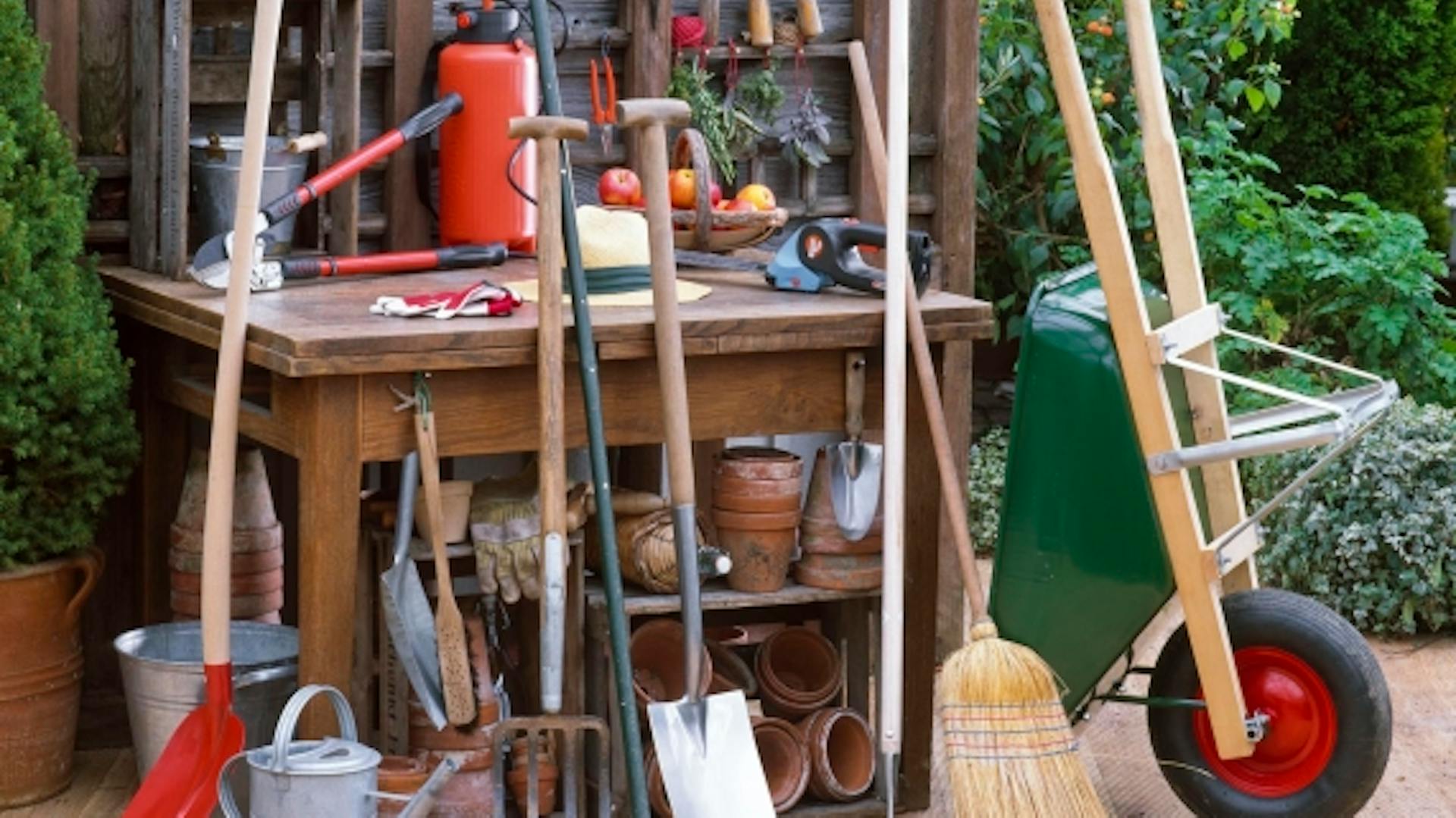 Raccolta di vari attrezzi da giardino davanti al banco da lavoro in legno, tra cui tagliasiepi, vanghe, scope e carriola.