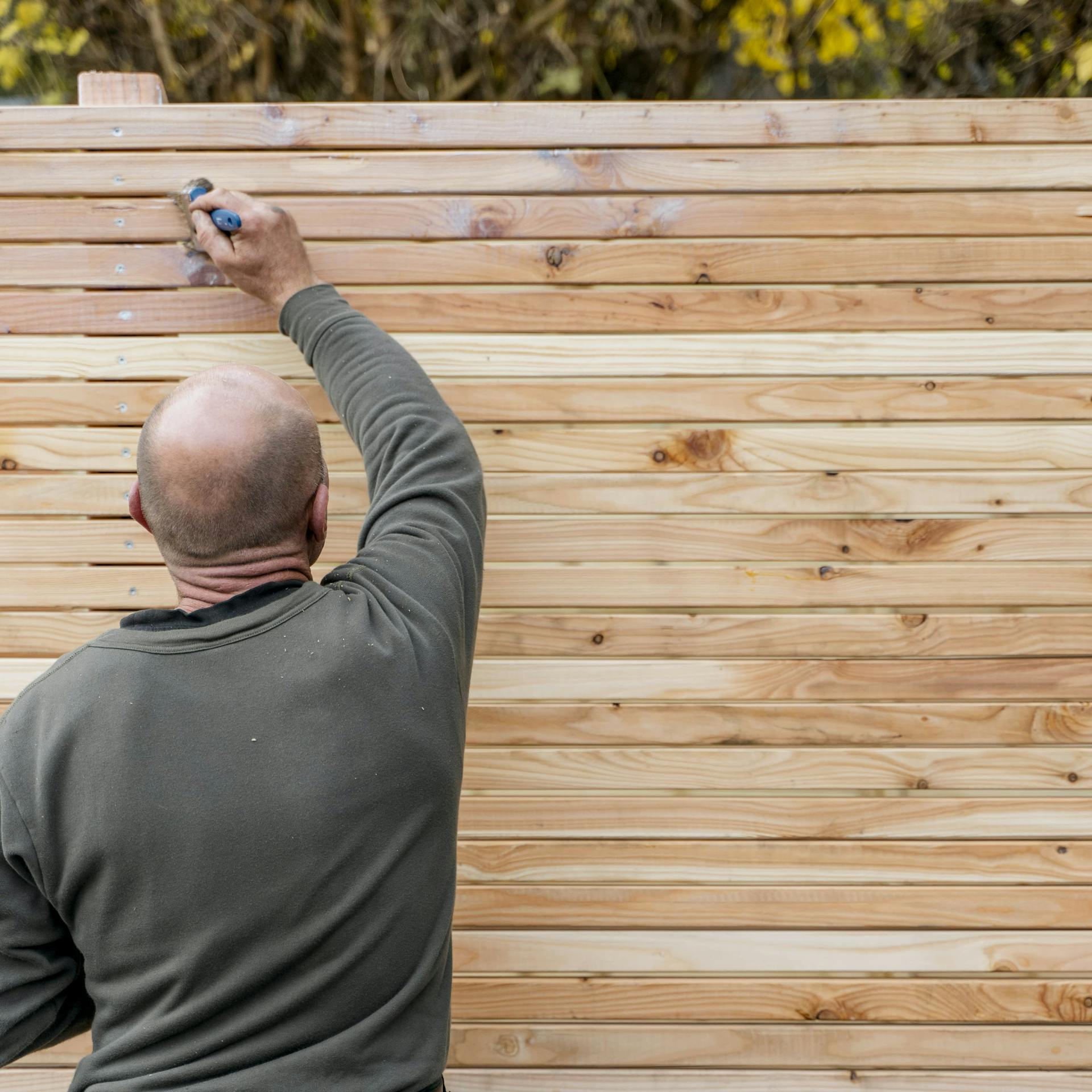 Ein Mann trägt Lasur auf den Sichtschutz aus Holz auf.