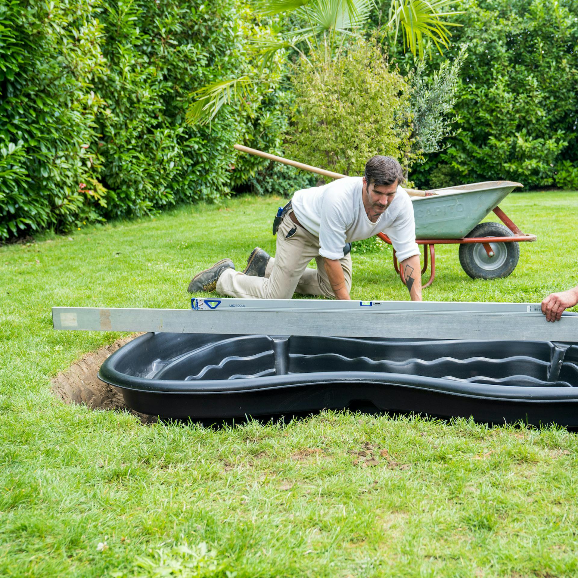 Heimwerker halten Wasserwaage auf über Teichbecken gelegte Richtlatte, um waagerechte Ausrichtung zu prüfen.