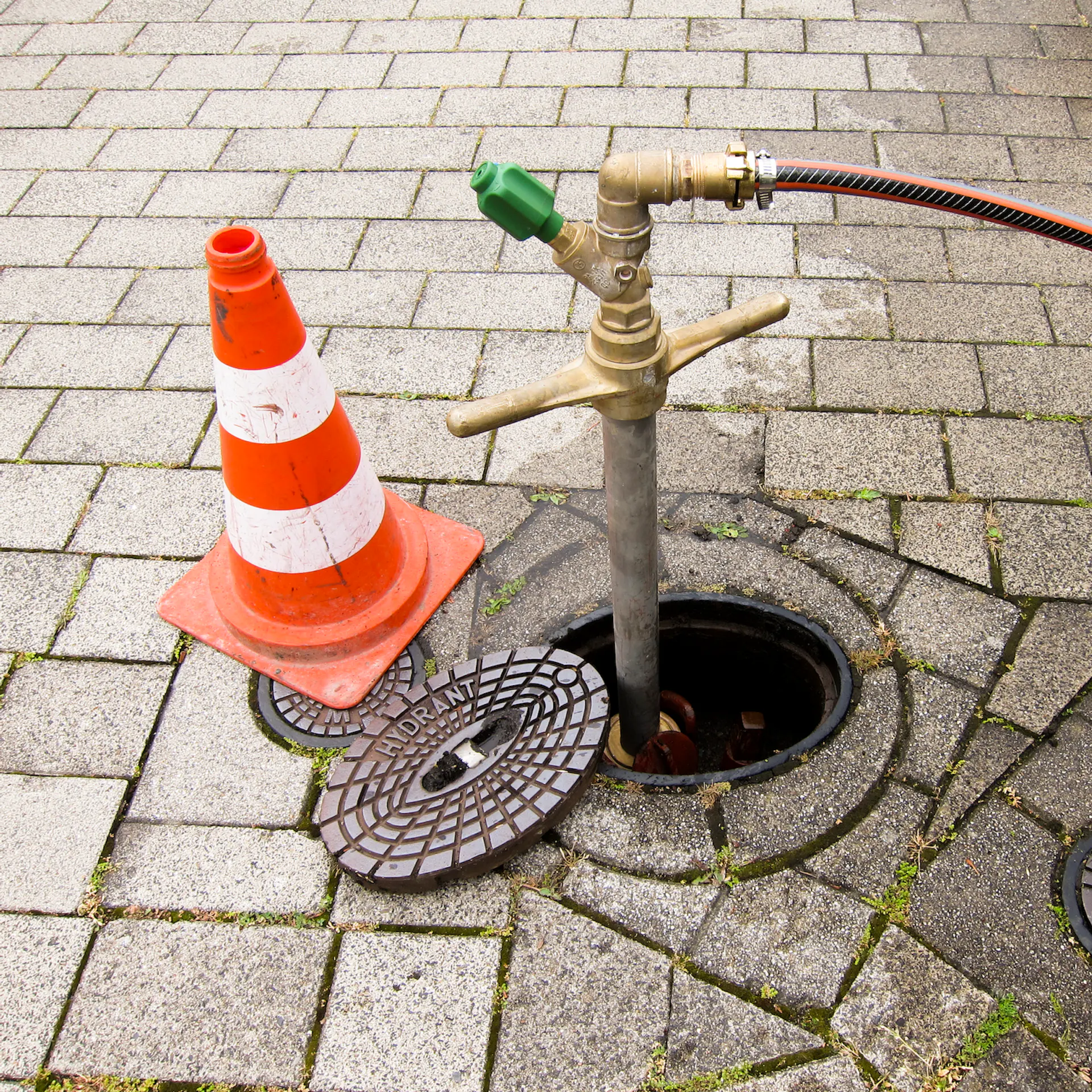 An einem Hydranten ist ein Gartenschlauch angeschlossen und links daneben steht ein Leitkegel.