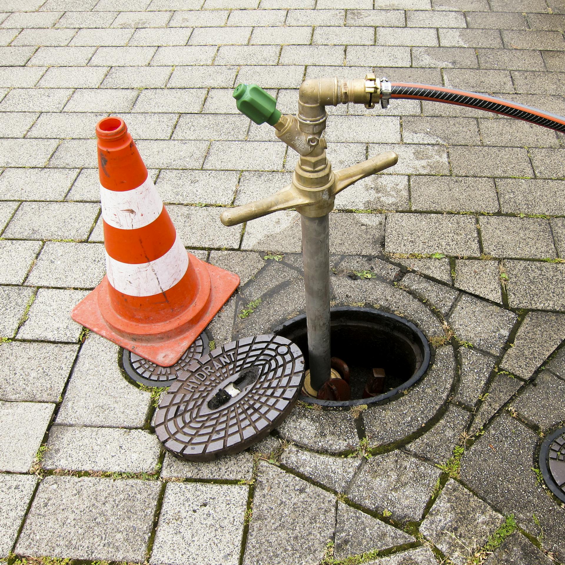 An einem Hydranten ist ein Gartenschlauch angeschlossen und links daneben steht ein Leitkegel.