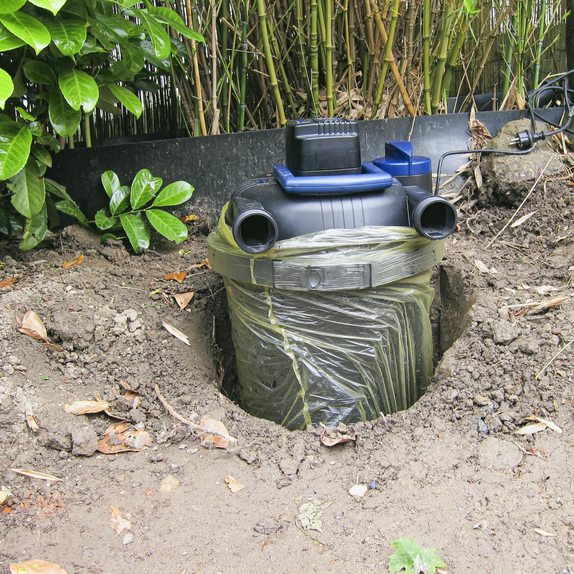 Um den Teichfilter ist eine Plastiktüte herumgebunden und er steckt in der Erde.
