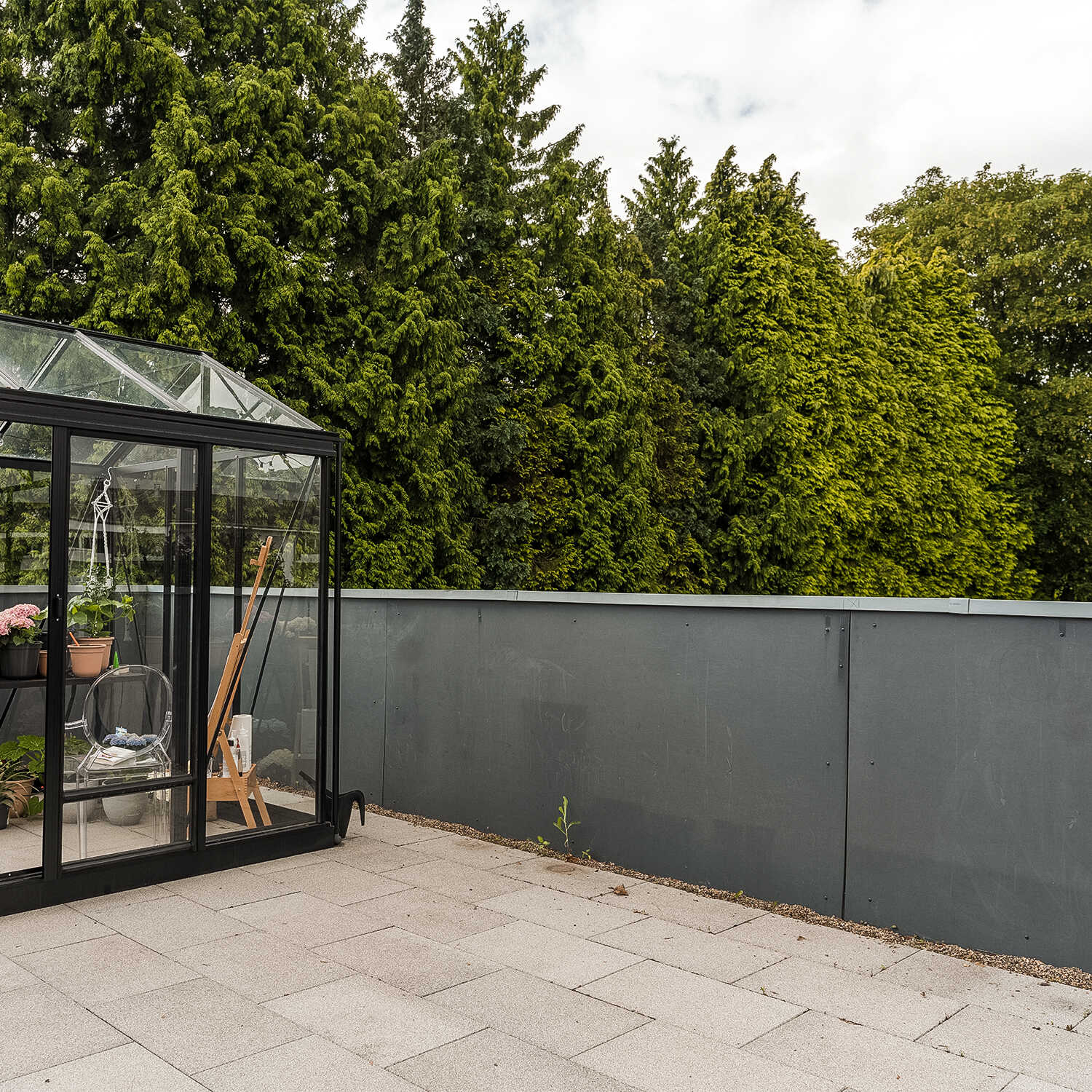 Karge Terrasse mit Gewächshaus und Bäumen im Hintergrund.