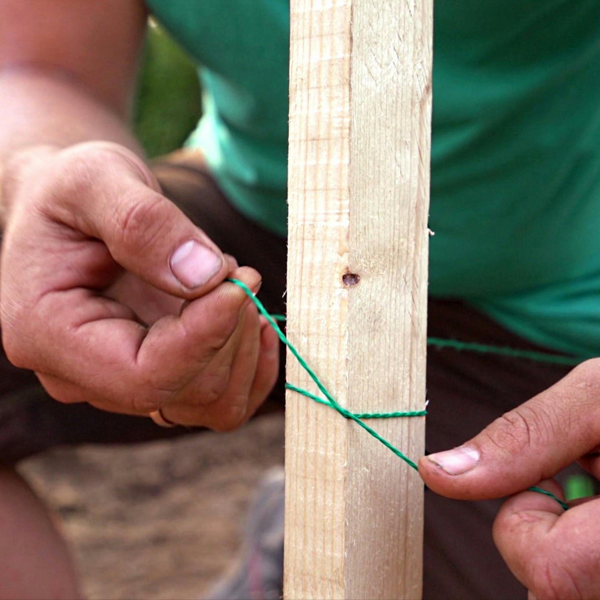 Foto zweier Hände, die die Schnur um einen Holzpfosten festziehen und so einen gestochenen Mastwurfknoten vollenden