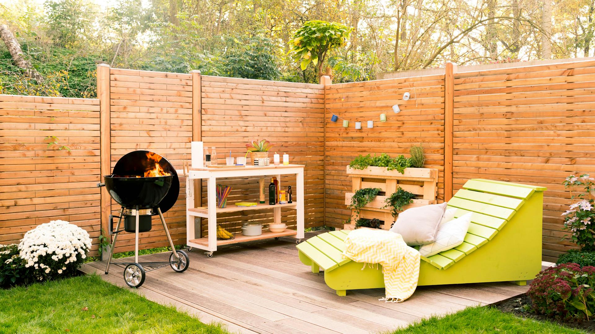 Terrasse aus Holz mit Grill, Gartenmöbeln und einem Gartenwagen sowie Pflanzregal und Sichtschutz aus Holz