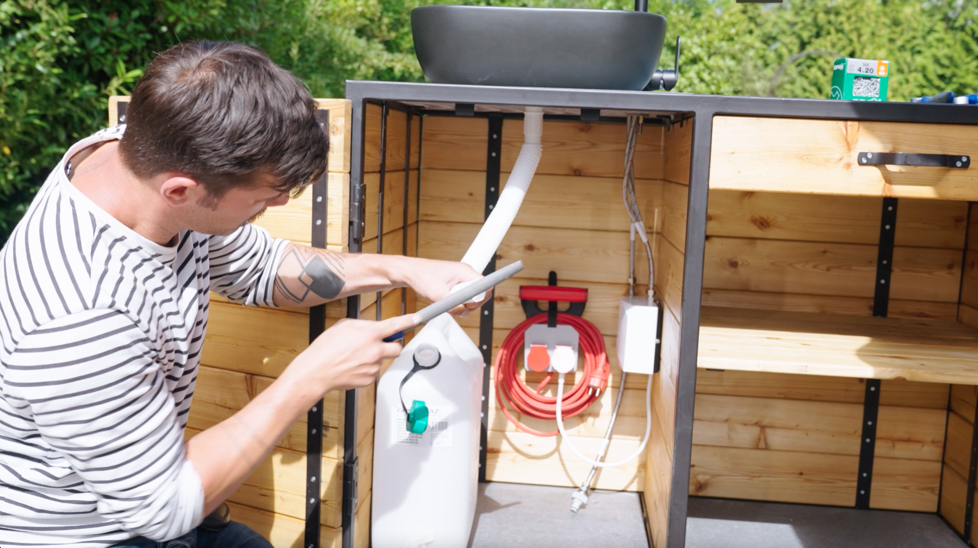 Base del lavandino in cucina esterna con serbatoio di scarico, scaldacqua istantaneo e presa elettrica. Un uomo accorcia un tubo che deve passare dallo scarico del lavandino al serbatoio delle acque reflue.