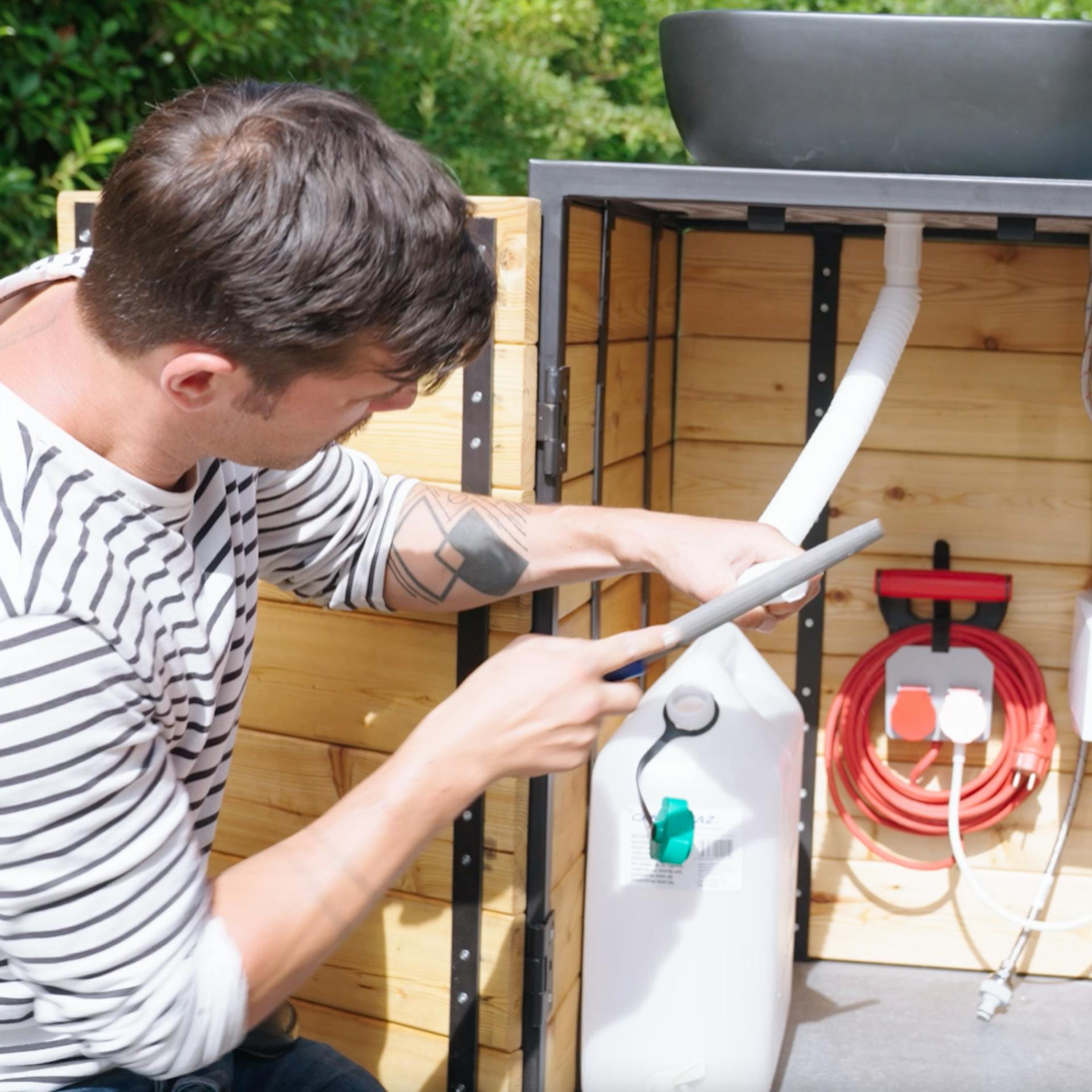 Base del lavandino in cucina esterna con serbatoio di scarico, scaldacqua istantaneo e presa elettrica. Un uomo accorcia un tubo che deve passare dallo scarico del lavandino al serbatoio delle acque reflue.