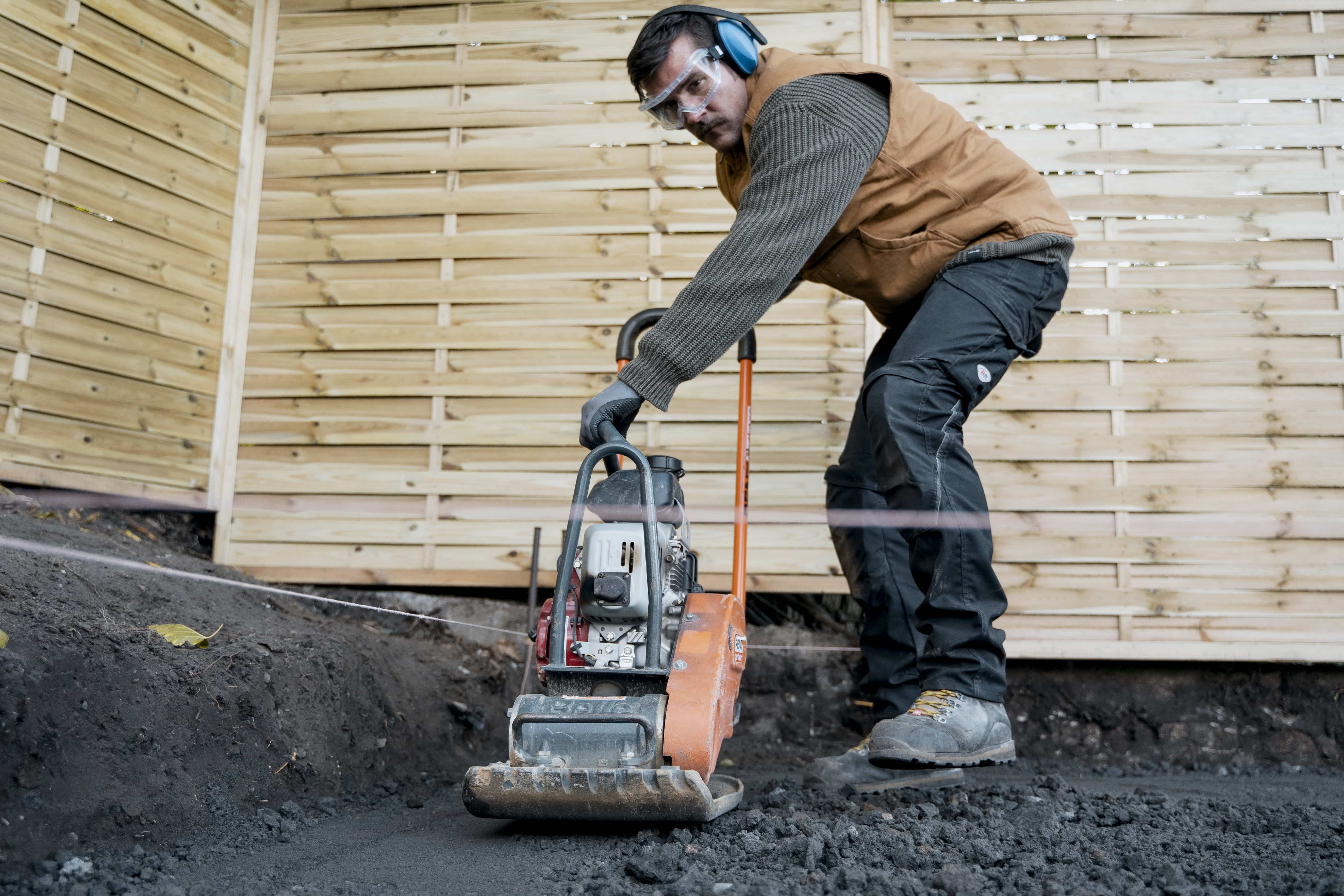 A hallásvédőt viselő munkás előtömöríti a kiásott területet egy lapvibrátorral..