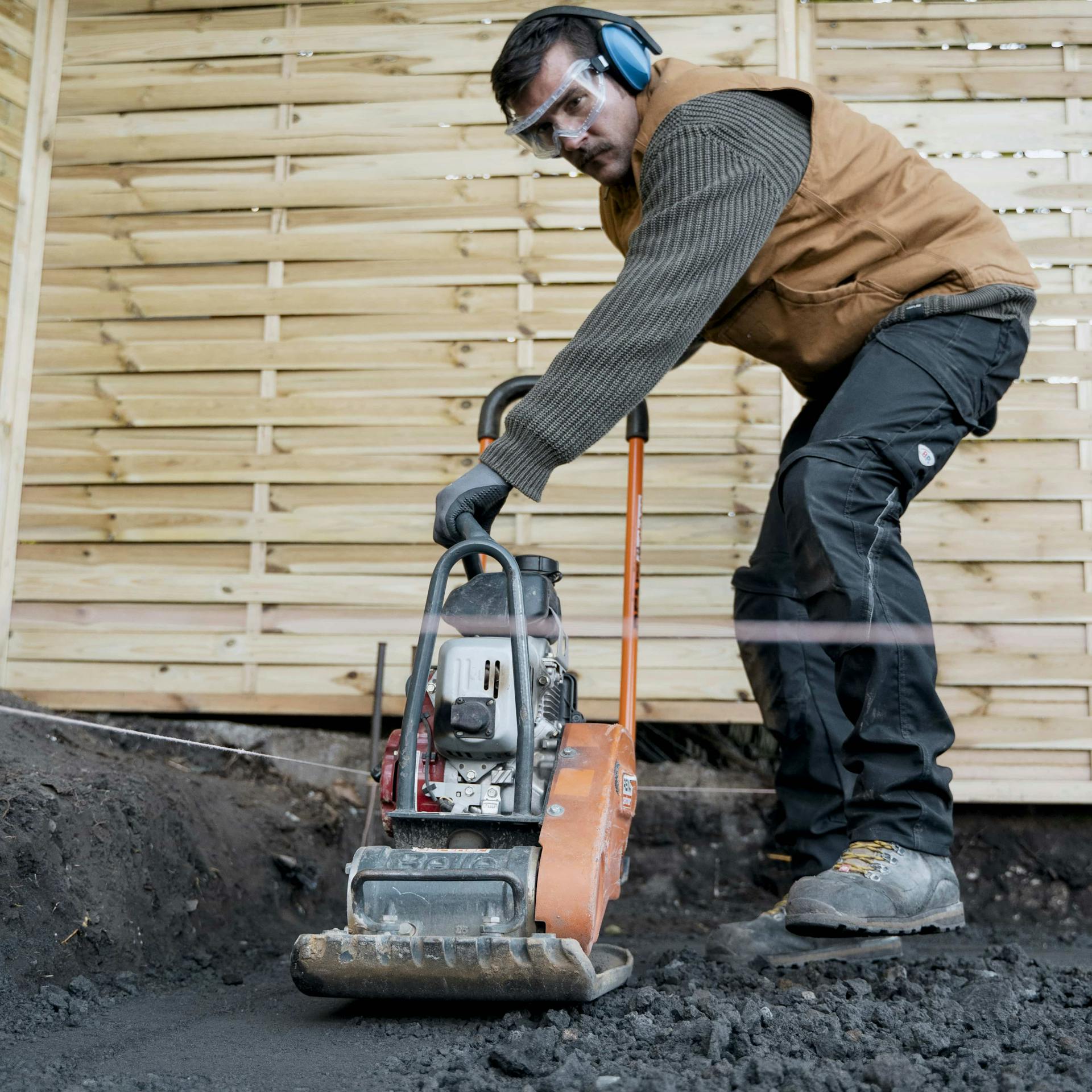 Handwerker mit Hörschutz verdichtet den ausgeschachteten Boden mit einer Vibrationsplatte vor.