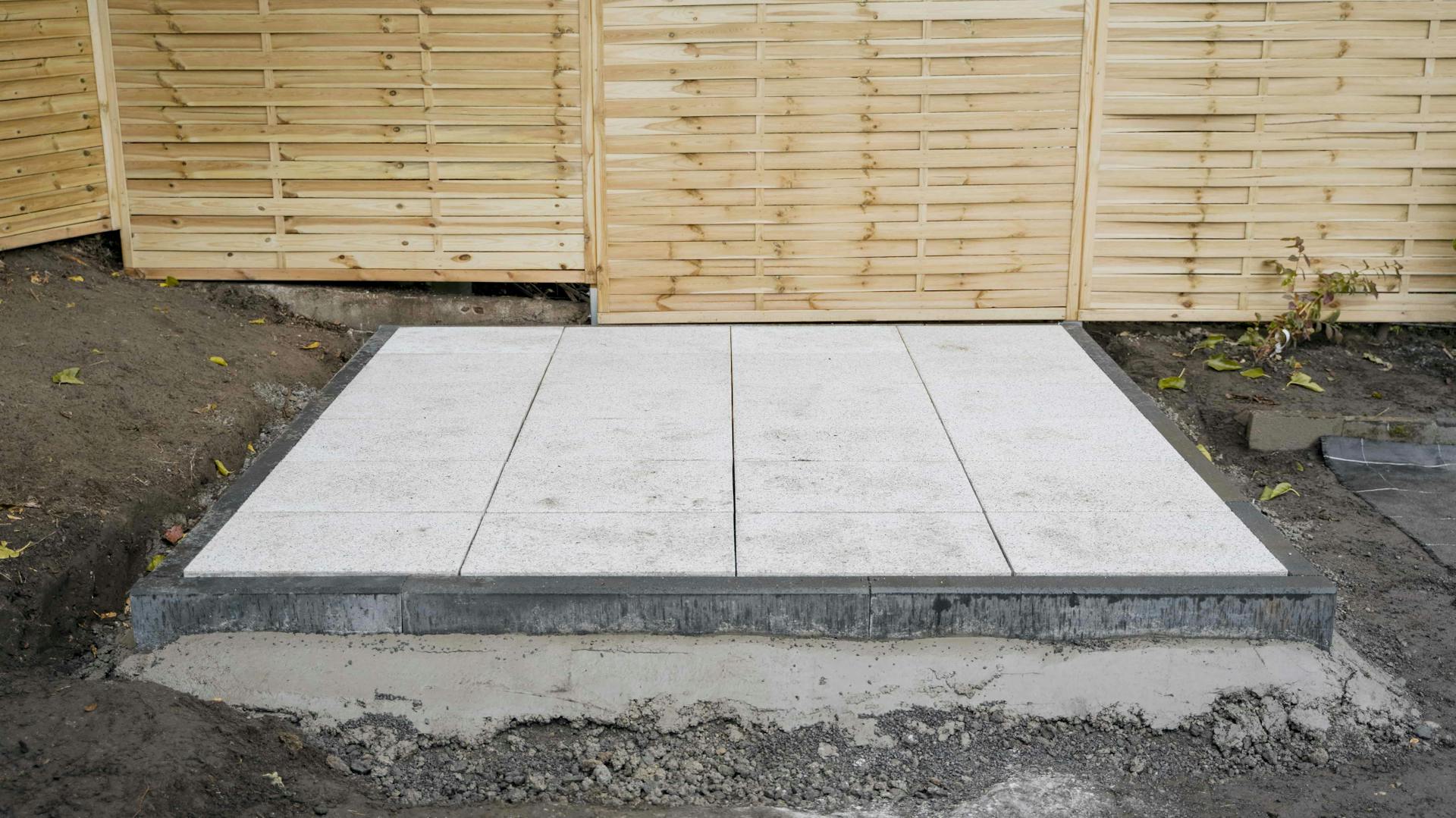 Vor einem Palisadenzaun aus hellem Holz sieht man ein fertiges Plattenfundament aus insgesamt 24 hellgrauen Gehwegplatten.