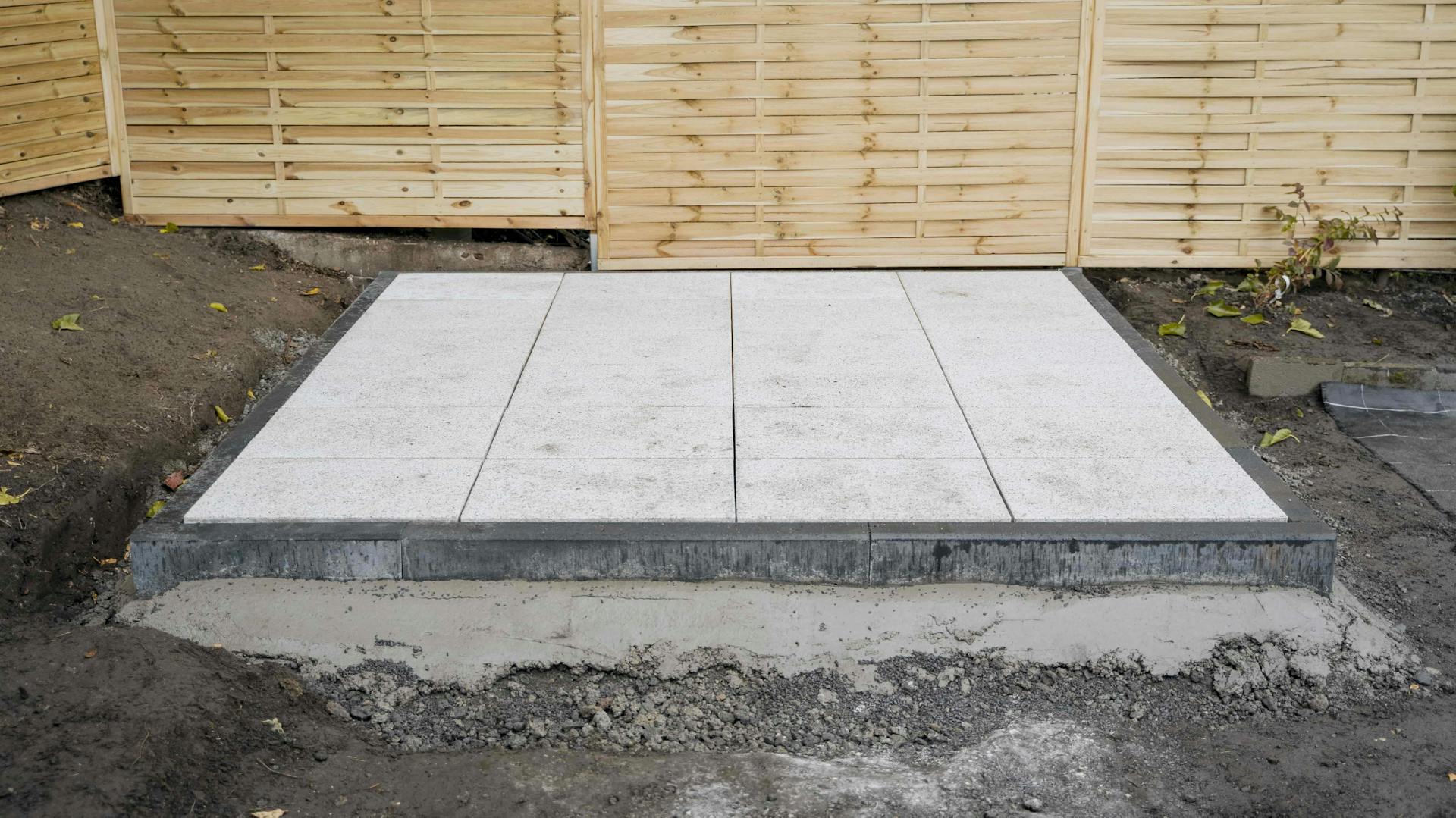 Vor einem Palisadenzaun aus hellem Holz sieht man ein fertiges Plattenfundament aus insgesamt 24 hellgrauen Gehwegplatten.