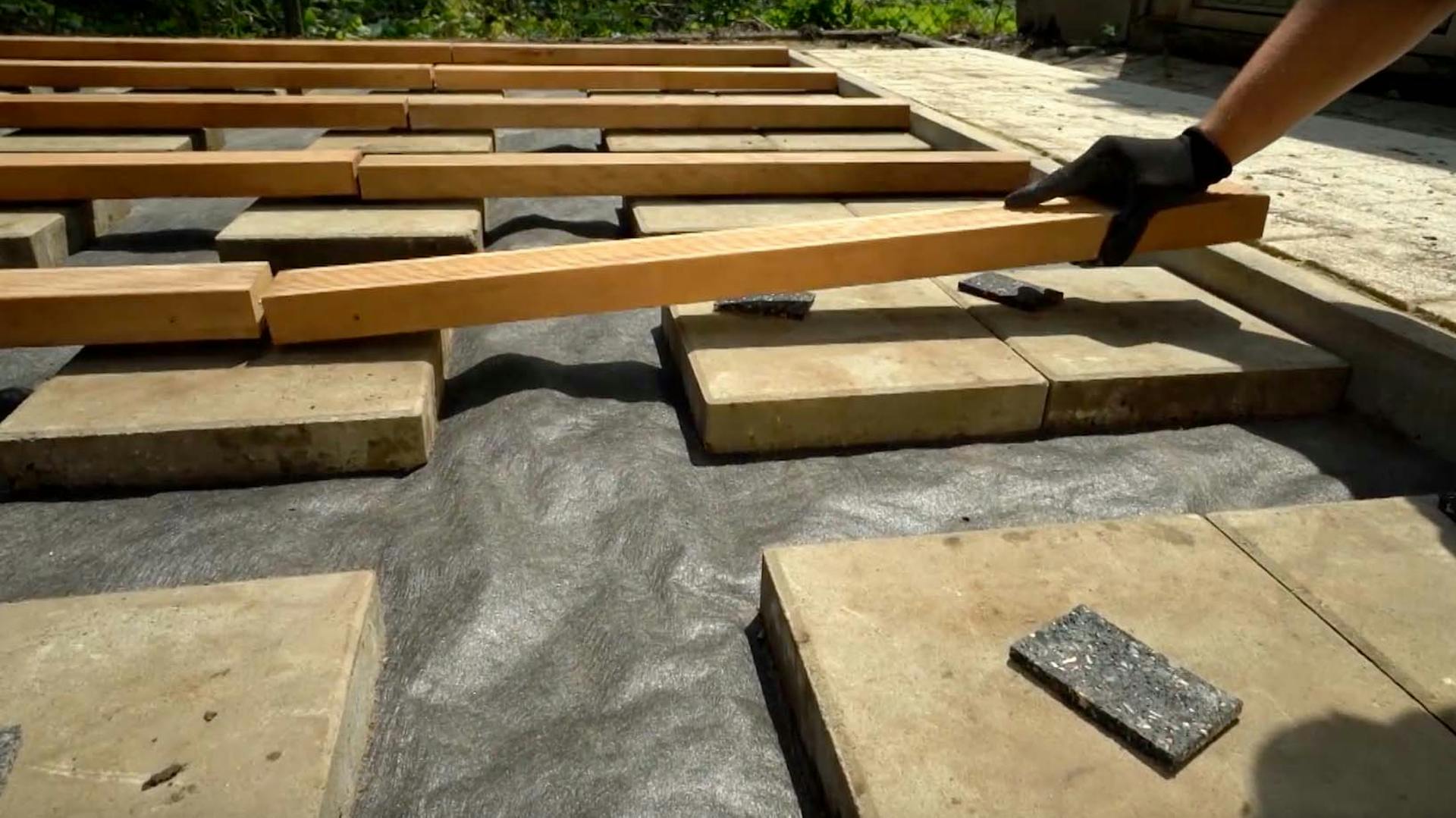 Un appassionato del fai-da-te posa le travi in legno della sottostruttura di una terrazza in legno su lastre di pietra con cuscinetti di appoggio.