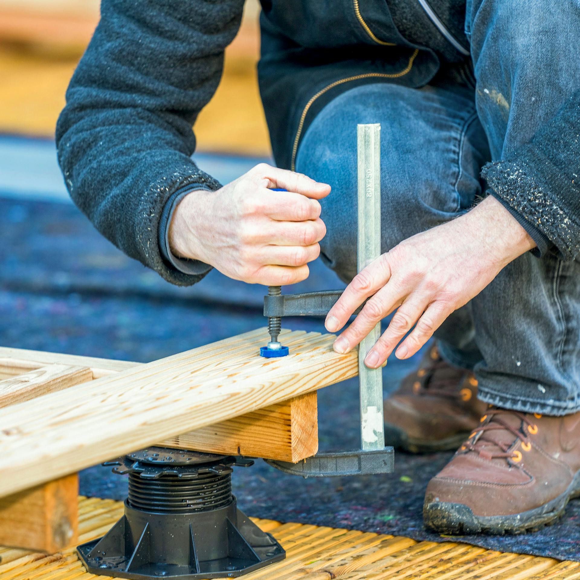 Hände befestigen eine Terrassendiele mit einer Schraubzwinge an einem Holzbalken.