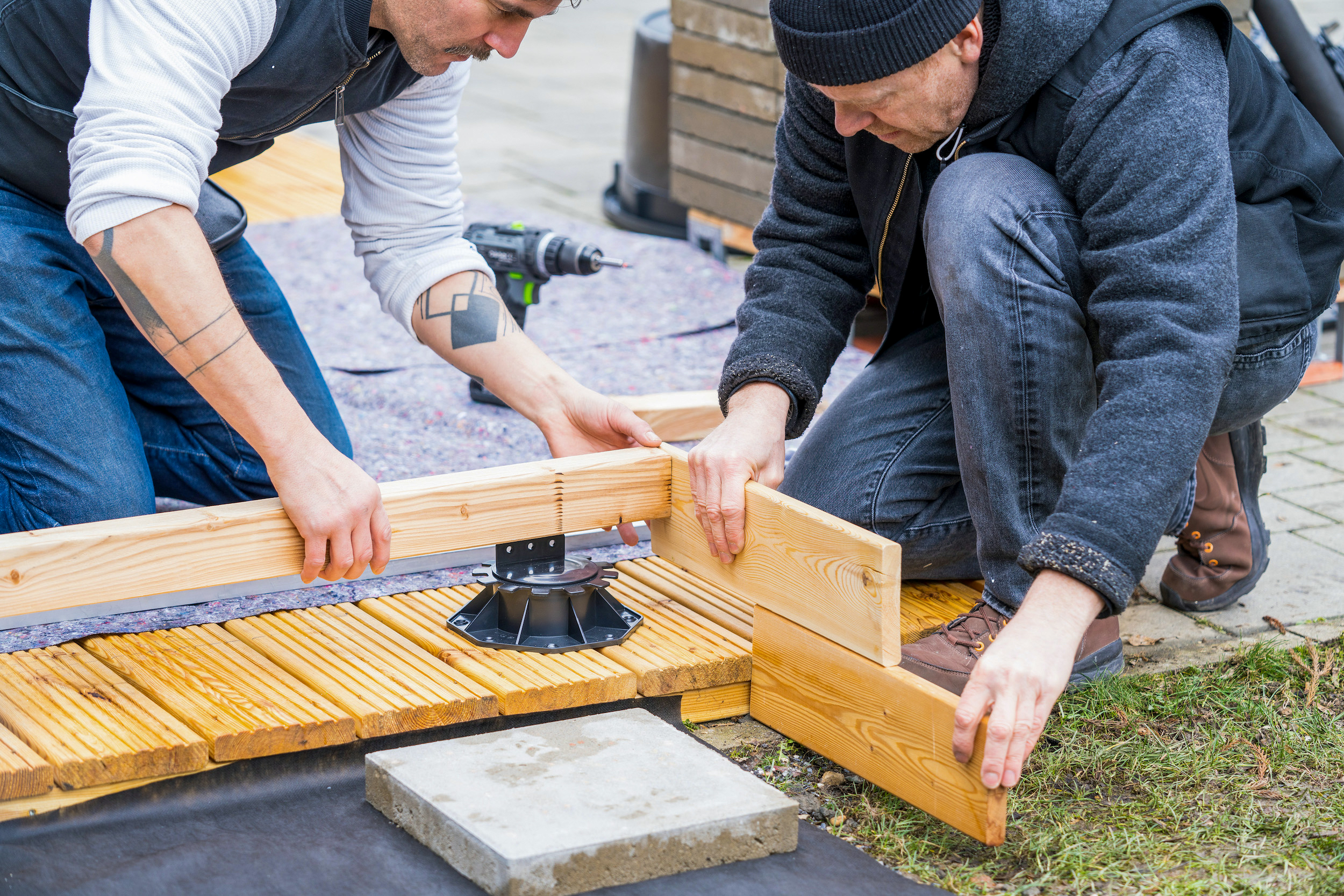 Due persone tengono delle assi di legno e una trave a un piedino.