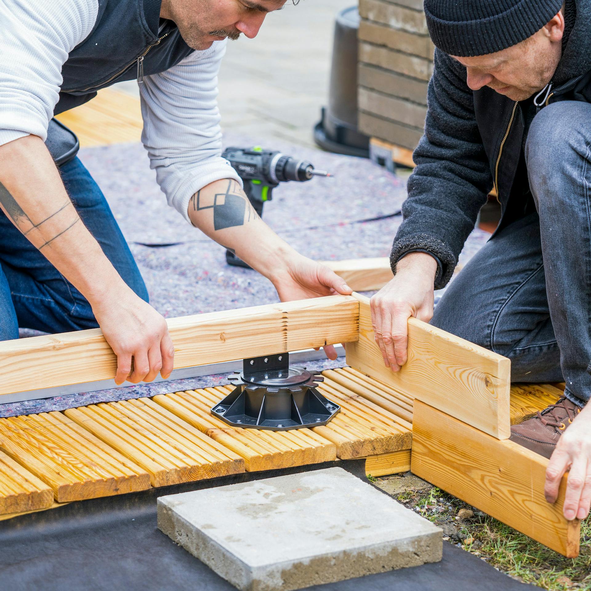 Zwei Männer halten Holzdielen und ein Kantholz an einen Stellfuß.