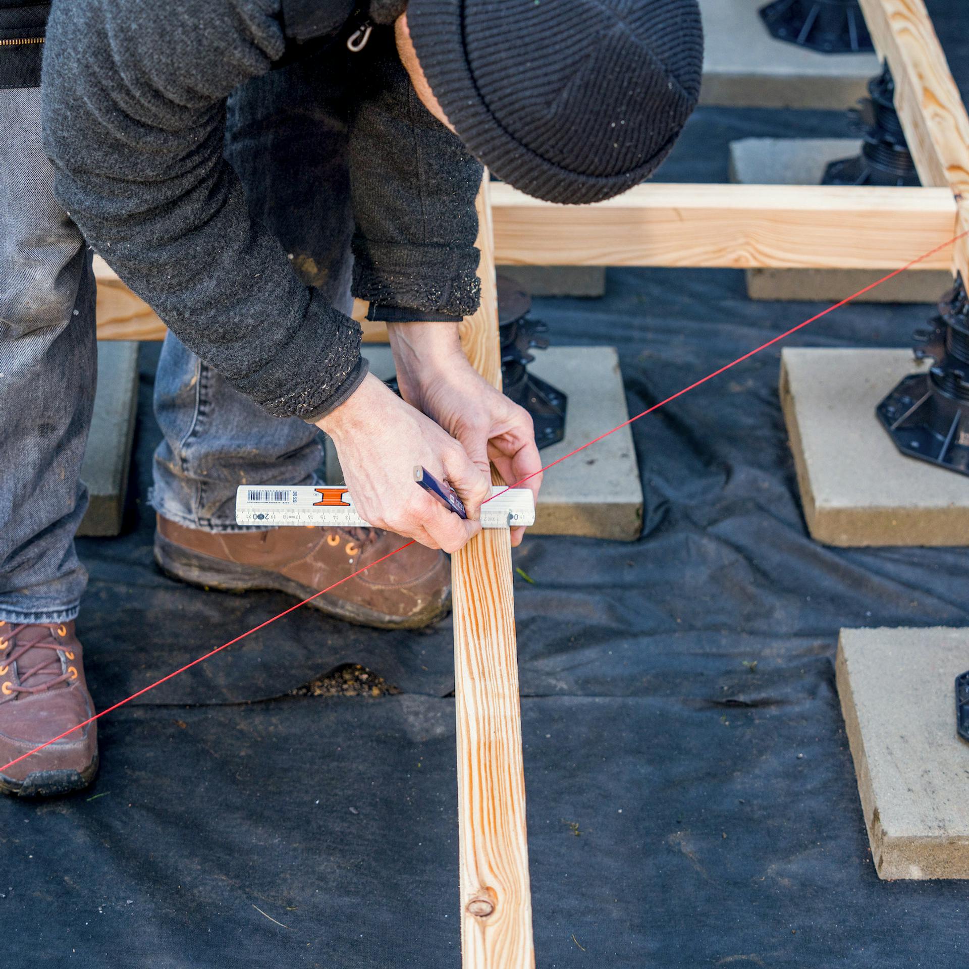 Ein Mann zeichnet mithilfe eines Schnurgerüsts eine Markierung auf einen Holzbalken.