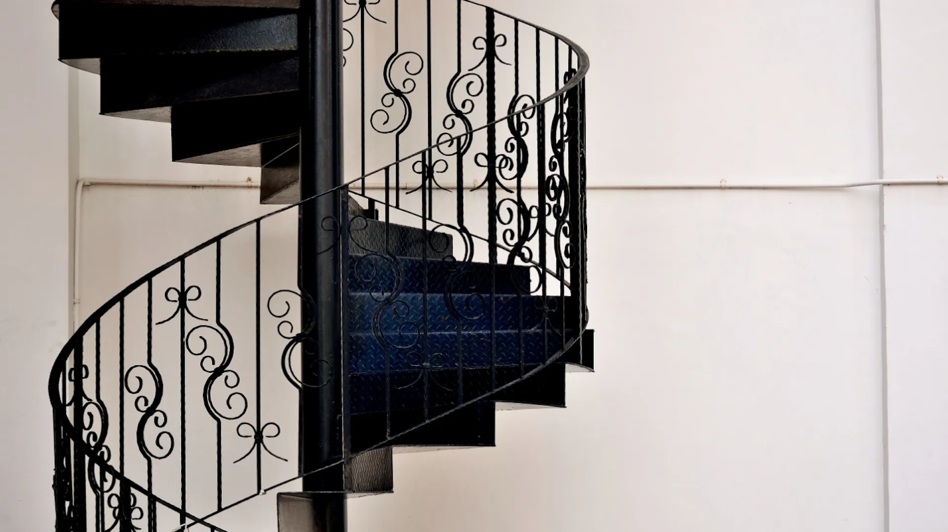 Nahaufnahme einer Wendeltreppe aus schwarzem Metall und verspieltem Geländer vor einem weißen Hintergrund.