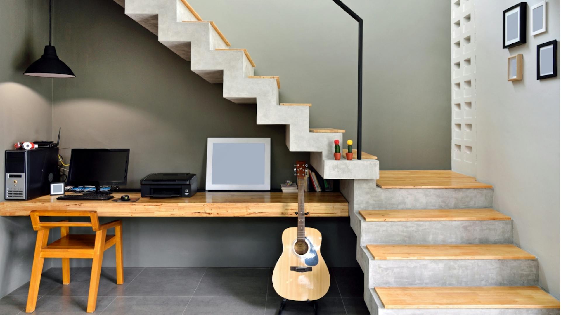 Es ist eine Treppe aus Beton zu sehen mit einem schlichten, schwarzen Geländer. Die Stufen sind mit Holz bedeckt. Unter der Treppe ist ein Schreibtisch zu sehen. Auf diesem stehen ein Computer sowie ein Bilderrahmen. Vor dem Tisch stehen ein Holzstuhl und eine Gitarre.