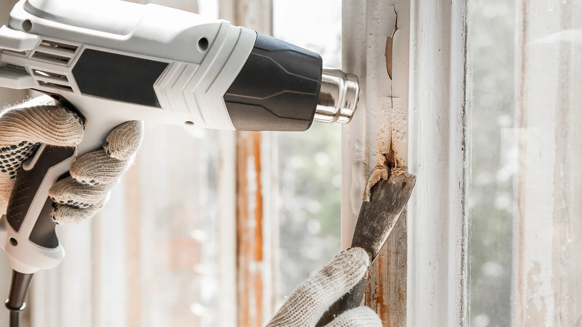 Heimwerker entfernt Latexfarbe mithilfe von Spachtel und Heißluftpistole vom Fensterrahmen.