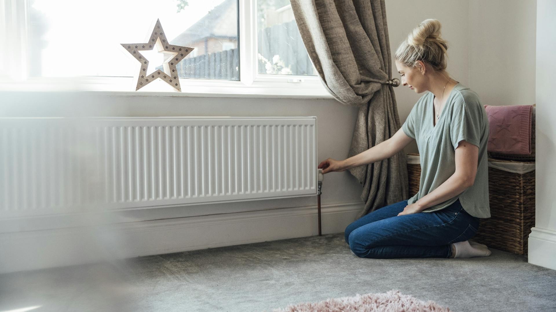 Egy ember ül egy nappaliban egy szőnyegen, radiátor mellett, és forgatja a termosztátot.