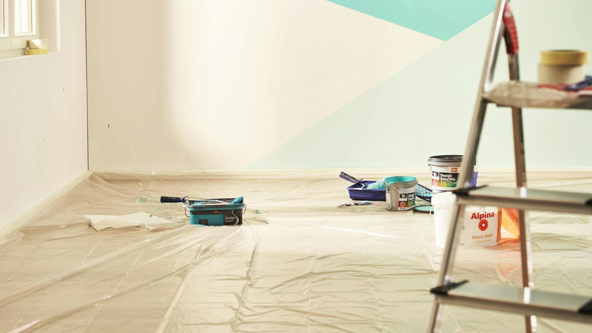 Wandgestaltung mit Blautönen in geometrischem Muster kurz nach dem Streichen mit folienbedecktem Boden, Pinsel, Farbe, Leiter und Malerkrepp