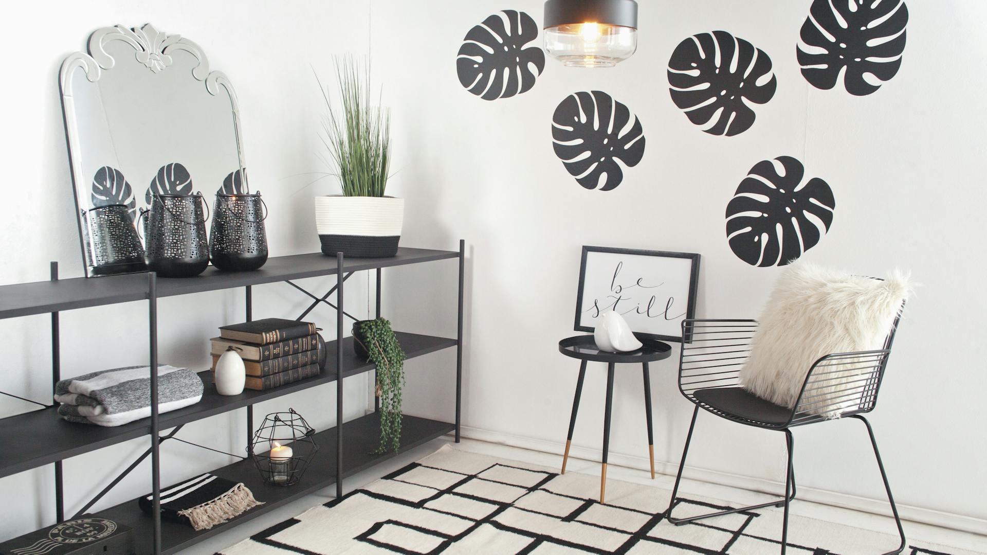 Ein Wandtattoo mit Silhouetten von Blättern in einem Raum, daneben stehen Stühle, auf der anderen Seite steht ein Regal mit einem Spiegel darauf.