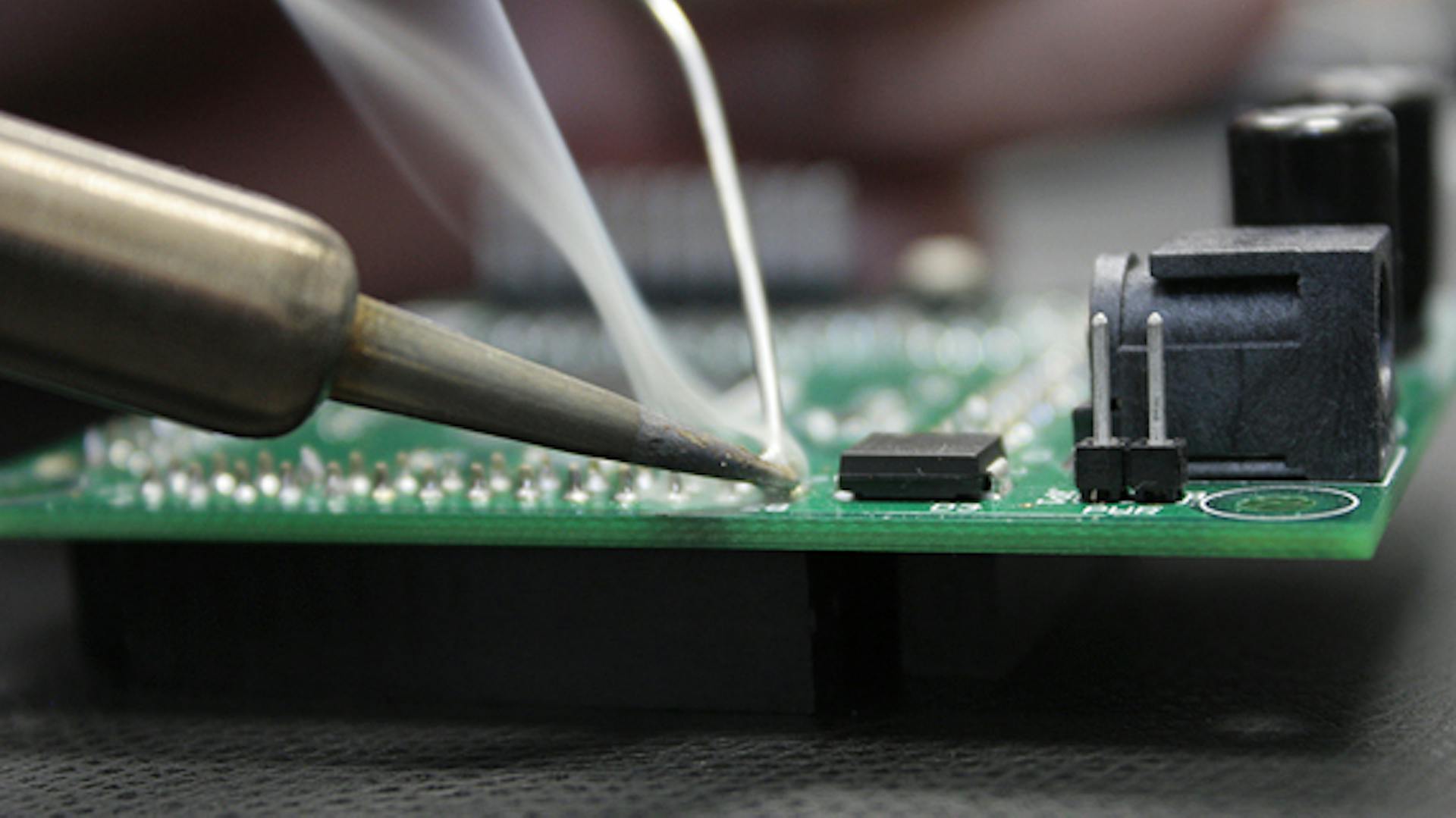 Ein Lötkolben bearbeitet eine Leiterplatte und ein Rauchfaden entsteht.