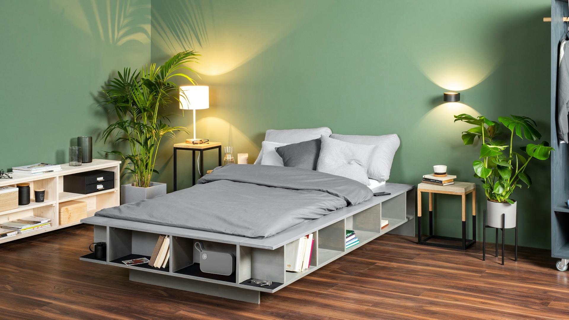 Graues Bett mit Regal im Rahmen auf dunklem Holzboden vor grüner Wand