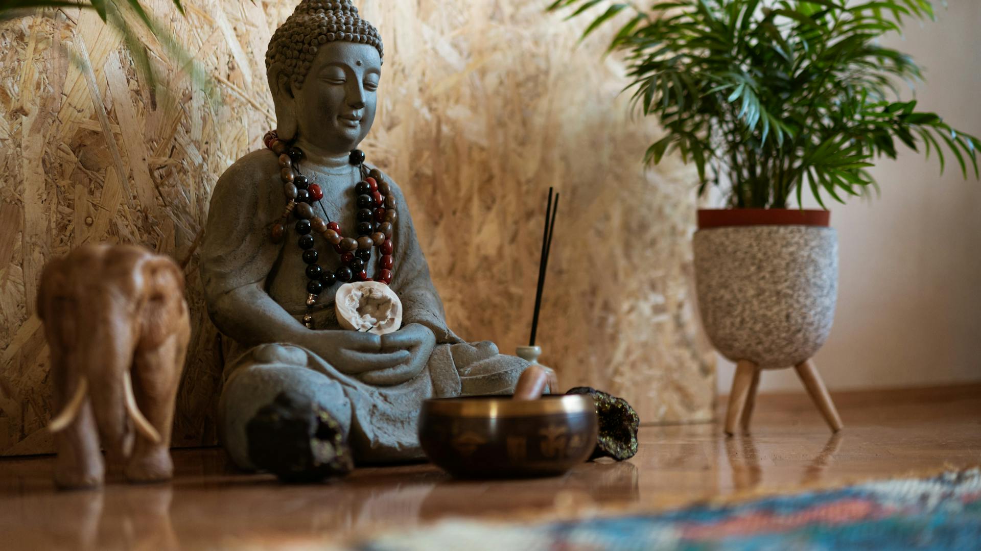 Ein Buddha mit Ketten umhangen sitzt auf dem Boden einer Wohnung. Neben ihm stehen ein Elefant und eine Pflanze.