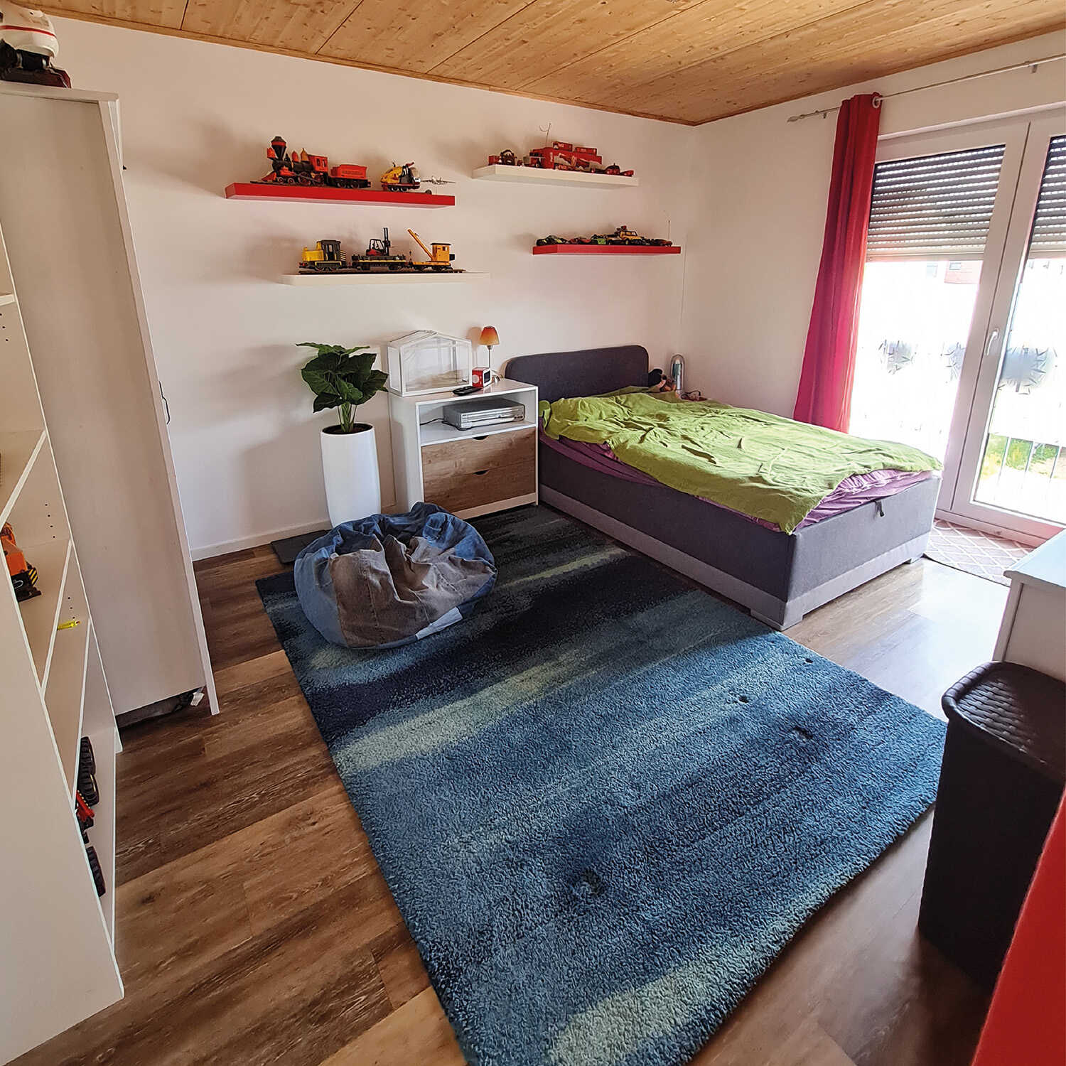 Ein Schlafzimmer in hellen Farben, mit einem großen, blauen Teppich in der Mitte, weißen Schränken und einem Bett in der Ecke des Zimmers.