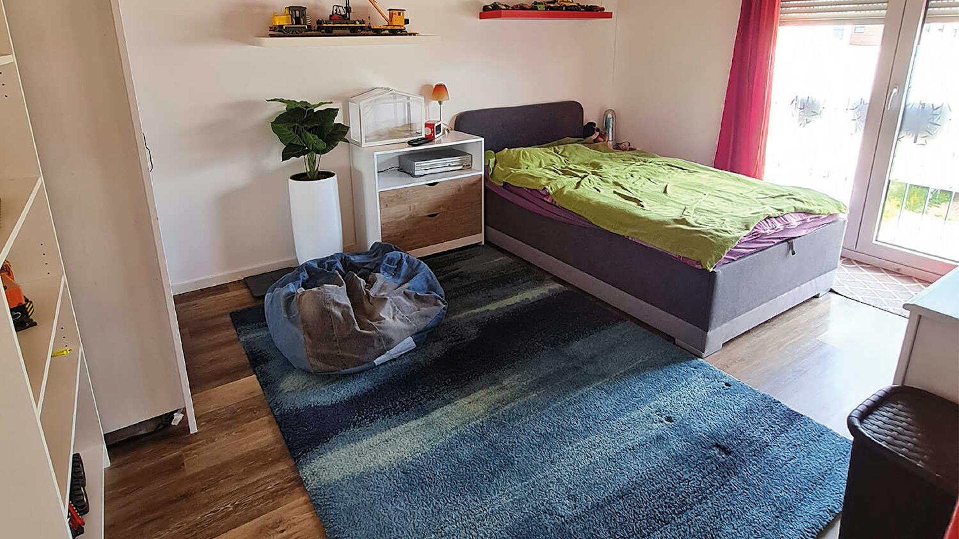 Ein Schlafzimmer in hellen Farben, mit einem großen, blauen Teppich in der Mitte, weißen Schränken und einem Bett in der Ecke des Zimmers.