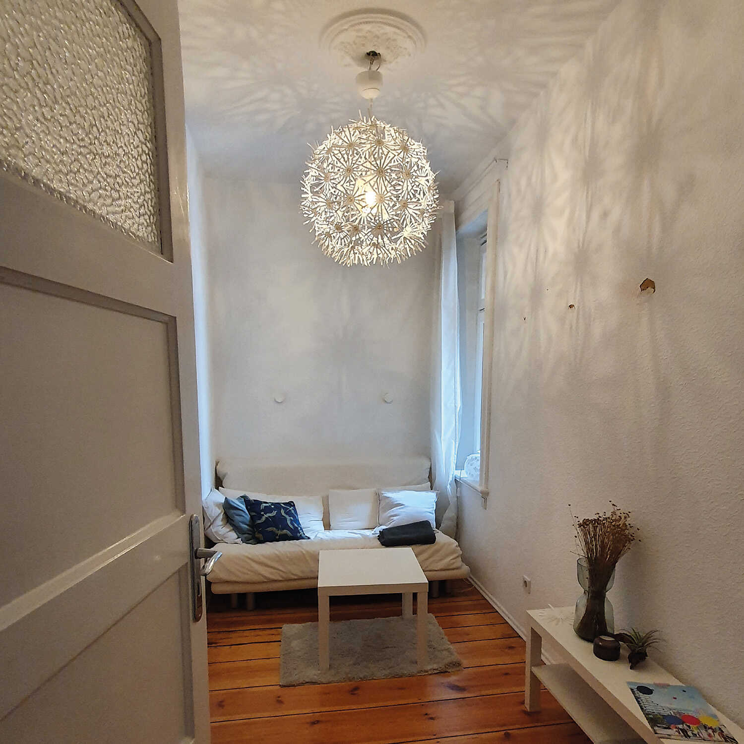In einem kleinen Raum, der weiß eingerichtet ist, steht an der hinteren Wand eine kleine Couch. Davor ist ein kleiner weißer Couchtisch platziert.