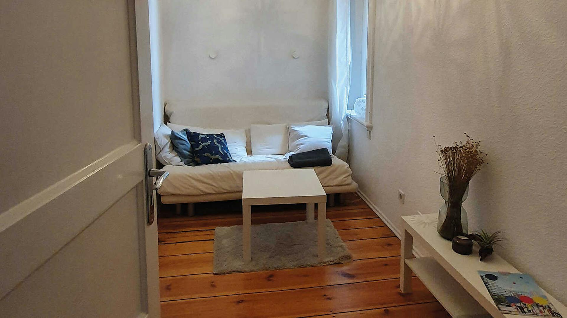 In einem kleinen Raum, der weiß eingerichtet ist, steht an der hinteren Wand eine kleine Couch. Davor ist ein kleiner weißer Couchtisch platziert.