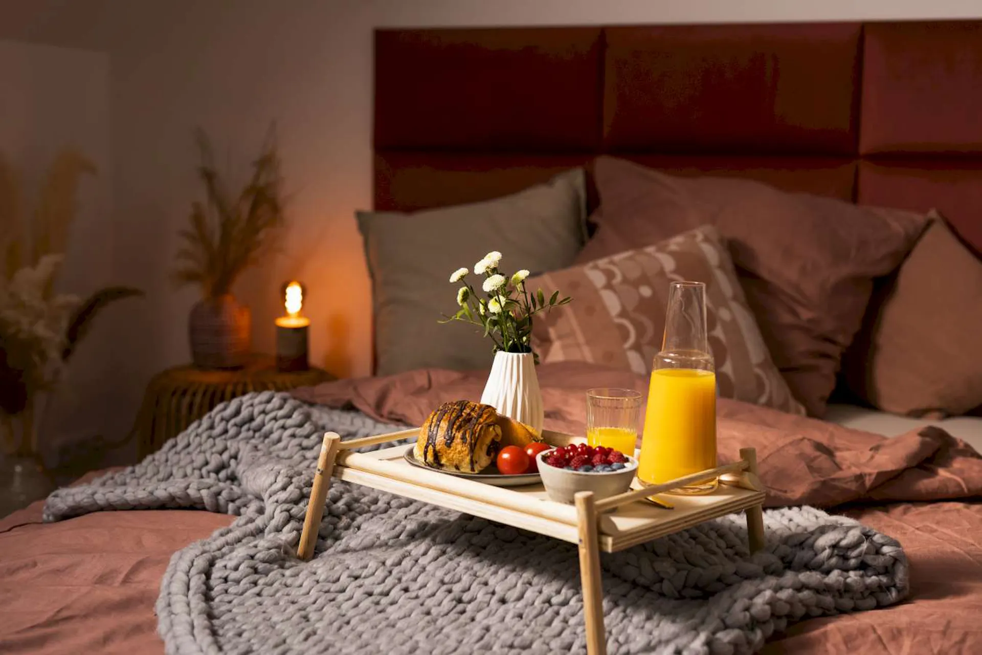 Ein selbstgebauter Tabletttisch, auf dem ein Croissant, Obst, Orangensaft und eine Vase mit Blumen steht.